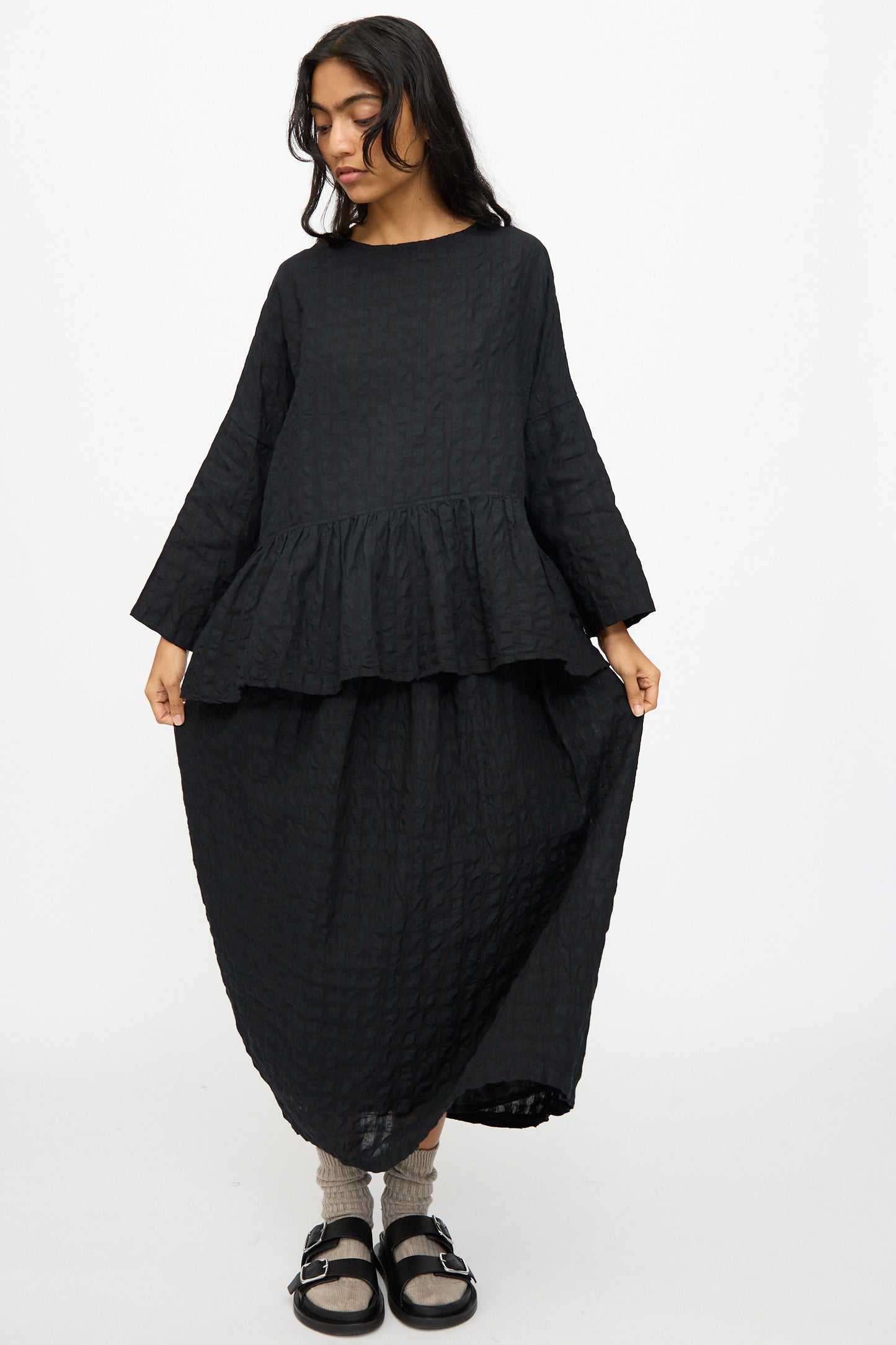 A person wearing an Ichi Antiquités Gingham Check Skirt in Black stands against a plain white background, paired with a textured black peplum top, grey socks, and black sandals.