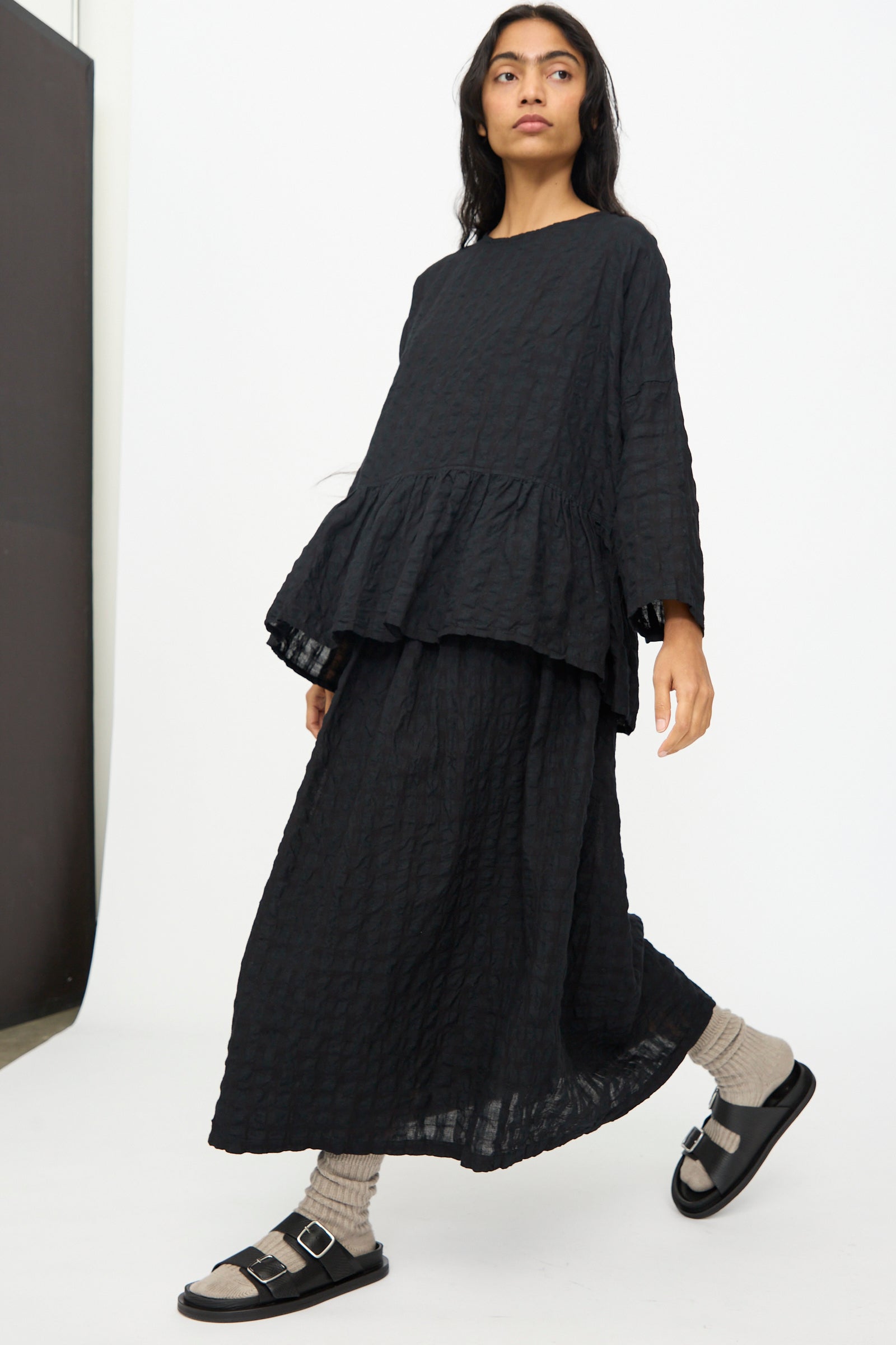 A woman wears the Ichi Antiquités Gingham Check Skirt in Black with a loose black top, gray socks, and black sandals as she walks through a minimalist space.