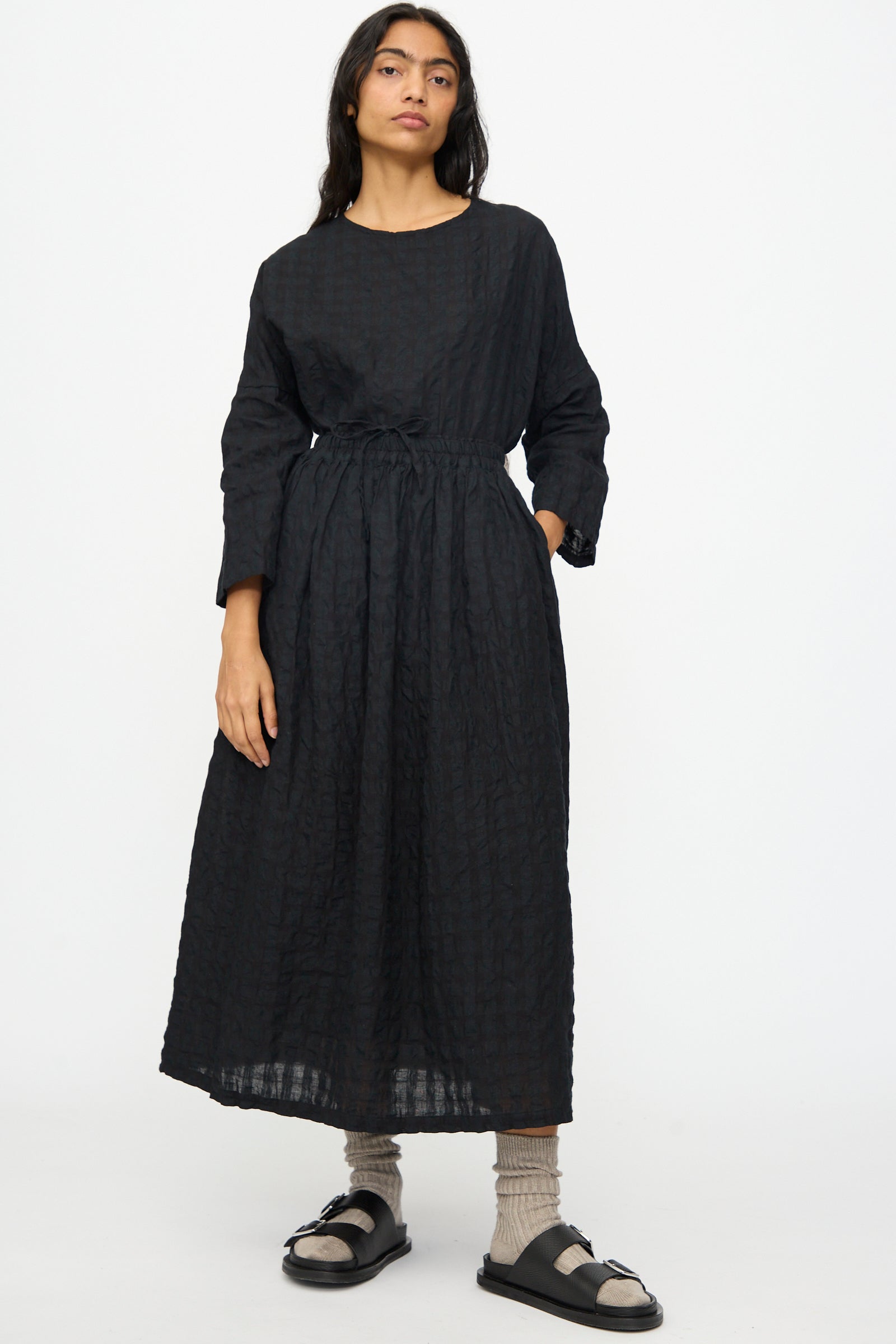 A person stands against a plain background, wearing the Ichi Antiquités Gingham Check Skirt in Black, paired with beige socks and black sandals.