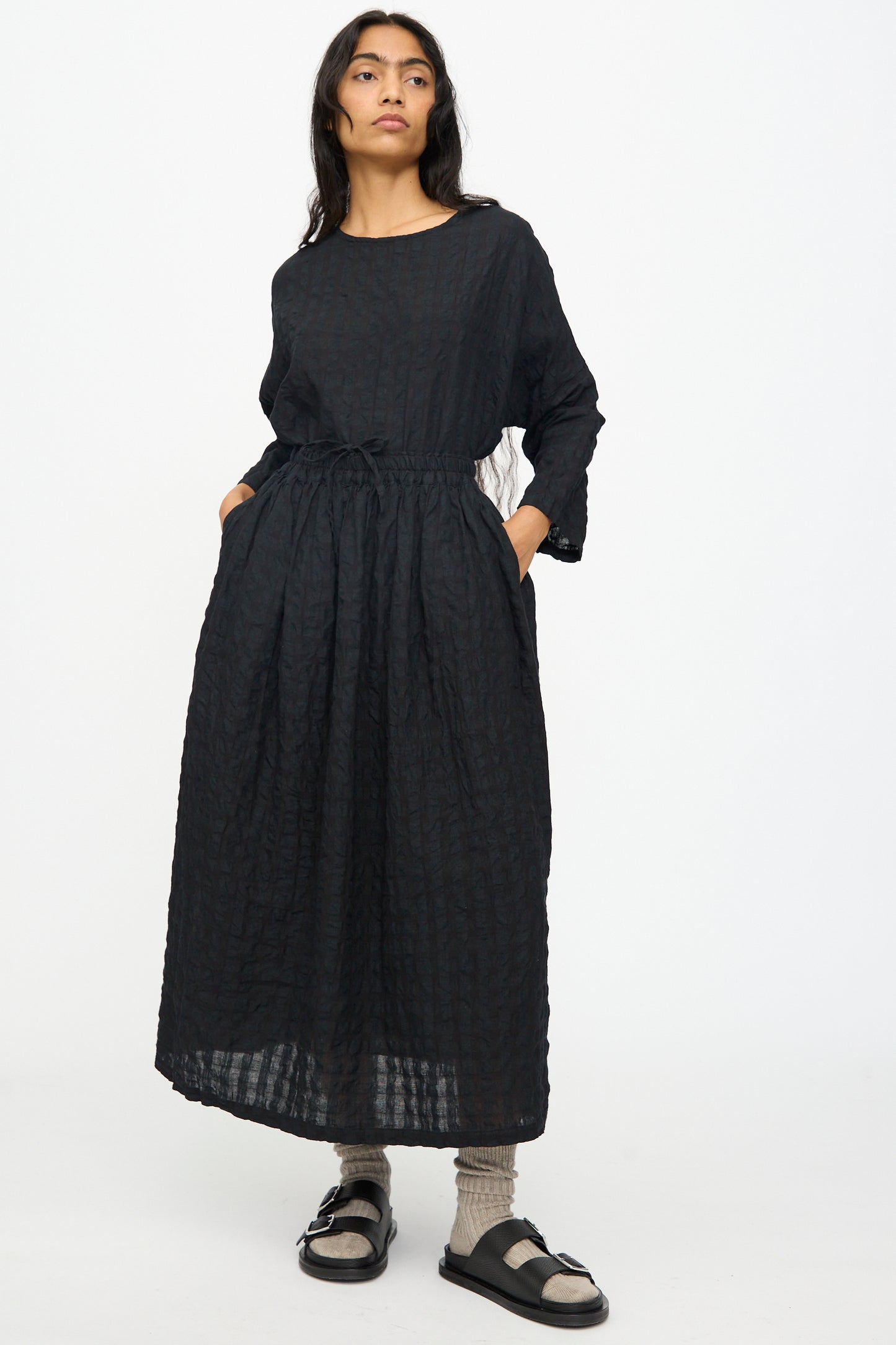 A woman stands with hands in pockets, looking to the side against a plain background, wearing the Ichi Antiquités Gingham Check Skirt in Black, paired with beige socks and black sandals.