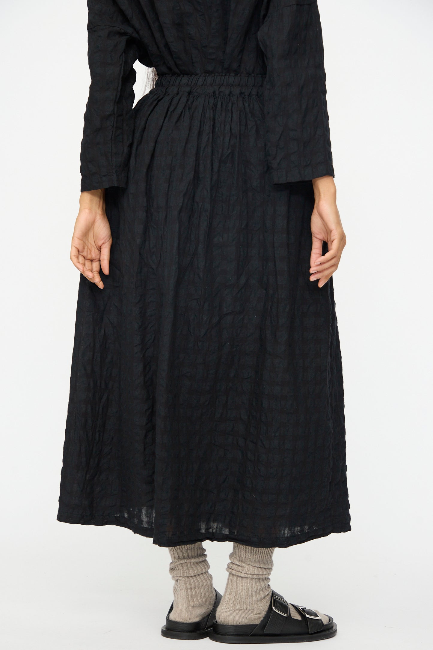 A person stands facing away, wearing the Ichi Antiquités Gingham Check Skirt in Black, paired with gray socks and black sandals against a plain background.