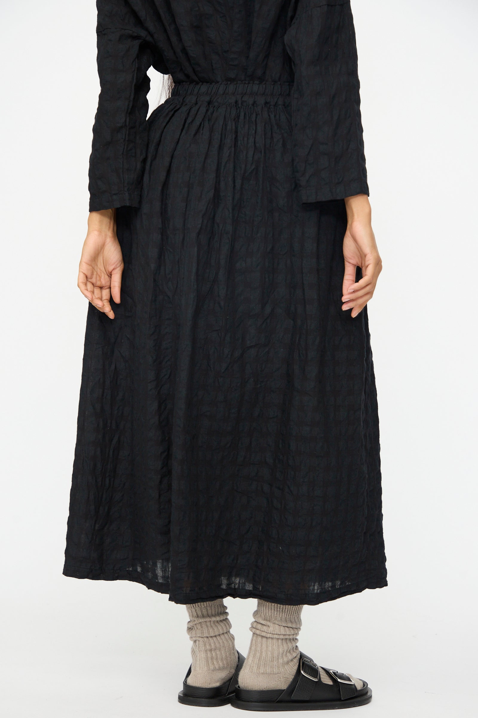 A person stands facing away, wearing the Ichi Antiquités Gingham Check Skirt in Black, paired with gray socks and black sandals against a plain background.