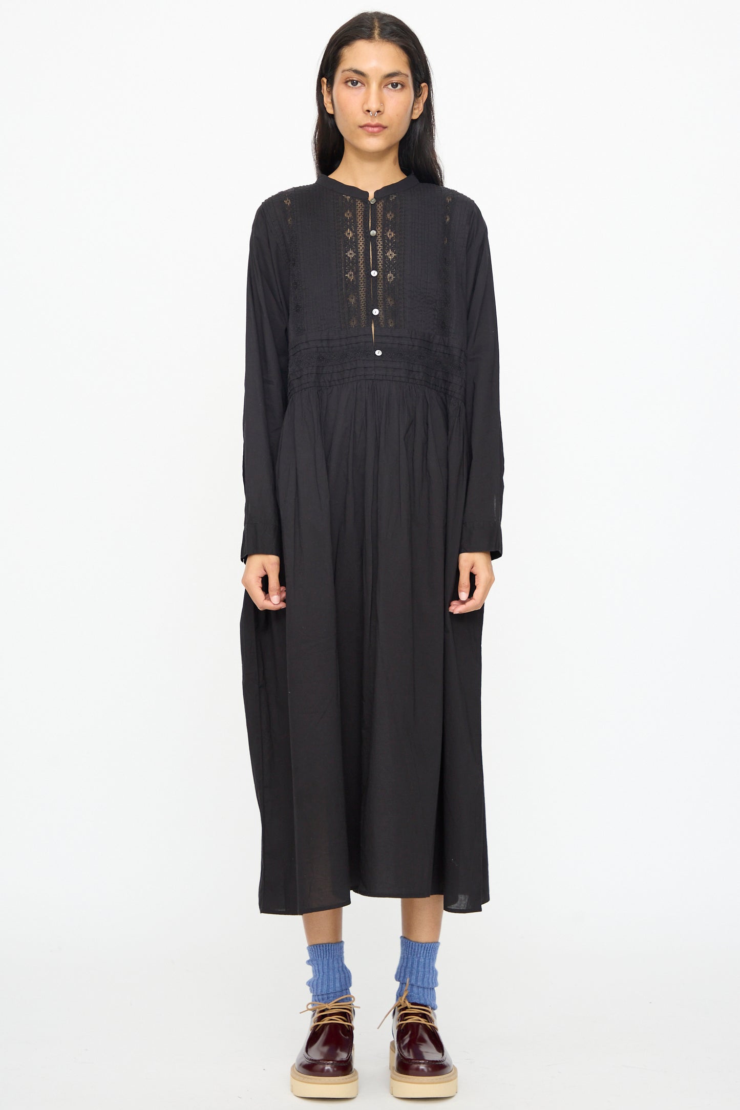 A person stands facing forward against a white background, wearing the Ichi Antiquités India Cotton Dress in Black with long sleeves, paired with blue socks and brown shoes.