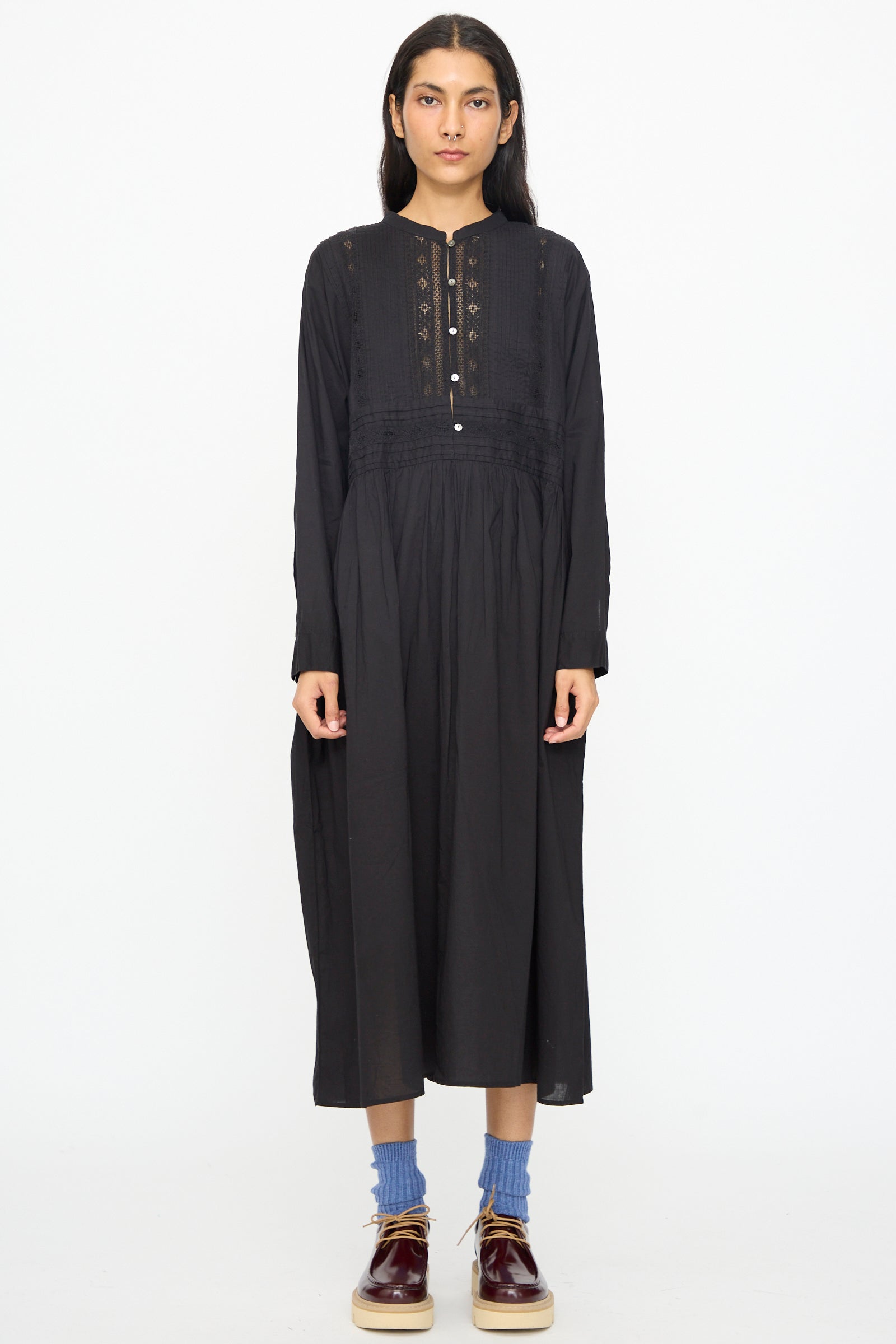 A person stands facing forward against a white background, wearing the Ichi Antiquités India Cotton Dress in Black with long sleeves, paired with blue socks and brown shoes.