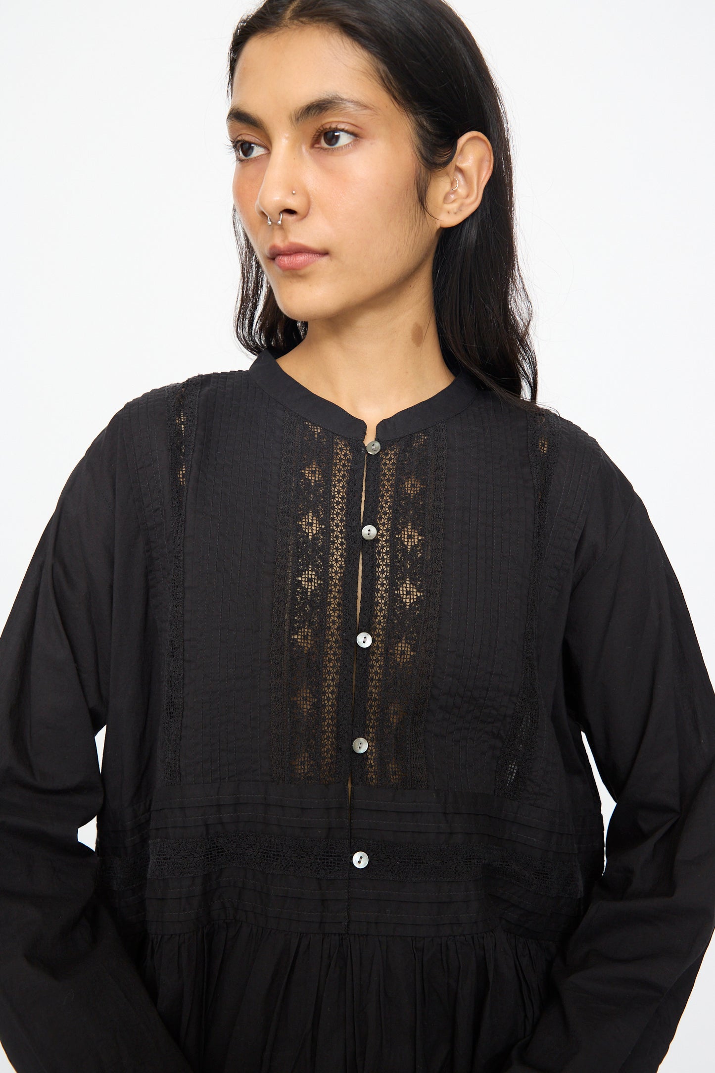 A person wears the India Cotton Dress in Black by Ichi Antiquités, featuring long sleeves, lace details, and a button front, while standing against a plain white background and looking slightly to the side.