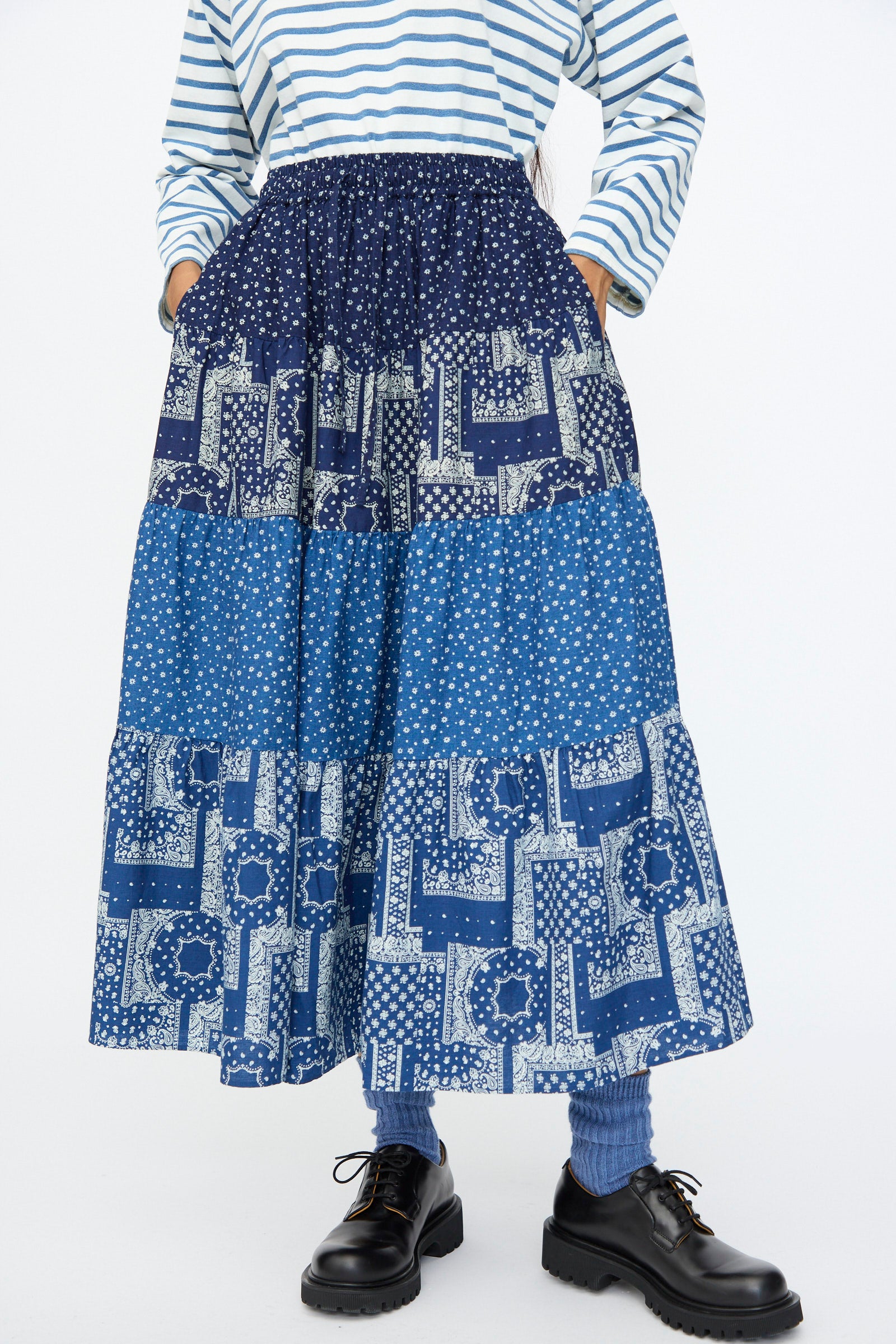 A person wearing a striped long-sleeve shirt, blue socks, black shoes, and the Indigo Flower Skirt in Navy Mix by Ichi Antiquités stands with hands in skirt pockets.