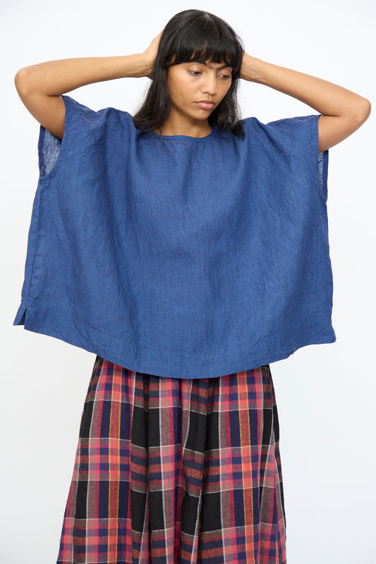 A person stands with hands behind head, wearing the Ichi Antiquités Indigo Linen Pullover in Blue and a red, black, and plaid skirt against a plain white background.
