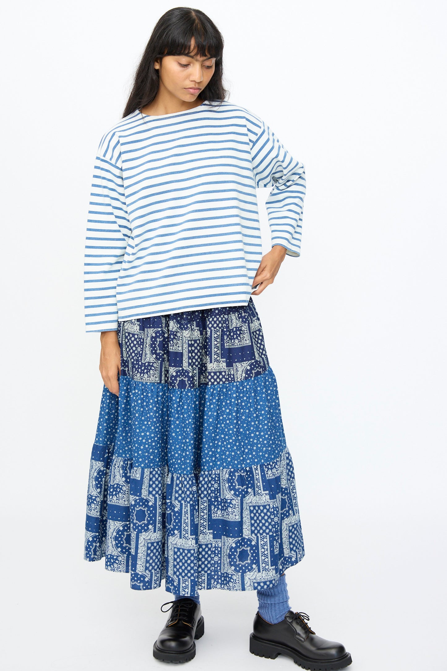 A woman wears the Ichi Antiquités Indigo Pullover Tee in White and Blue, paired with a blue patterned maxi skirt, blue socks, and black shoes, standing against a plain white background.