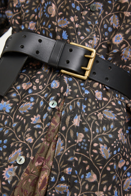 A black Ichi Antiquités leather belt with a brass buckle fastened over a floral print shirt and brown trousers.