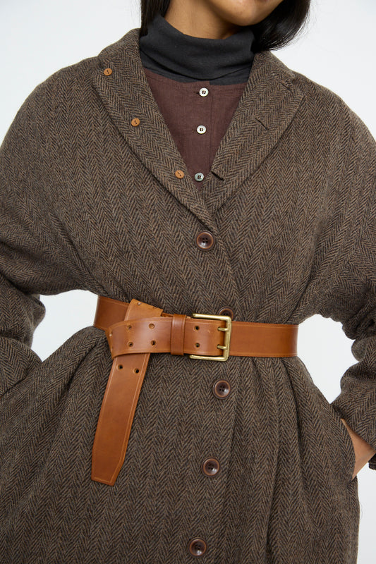 A person wears a herringbone pattern brown coat, buttoned down and cinched at the waist with Ichi Antiquités' Leather Belt in Brown featuring a gleaming brass buckle. A black turtleneck and brown shirt peek out beneath.