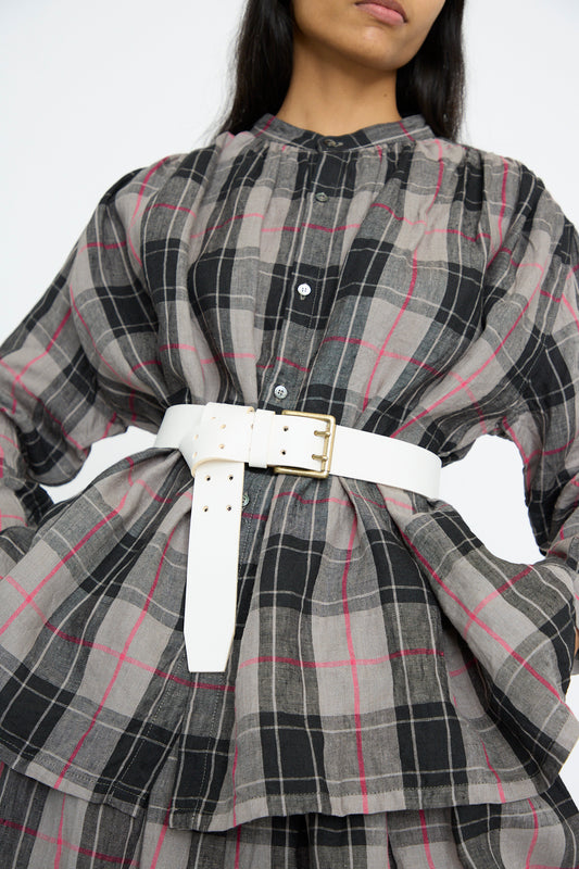 A person wearing a plaid shirt dress is styled with the Ichi Antiquités Leather Belt in White, featuring a brass buckle, against a white background. 