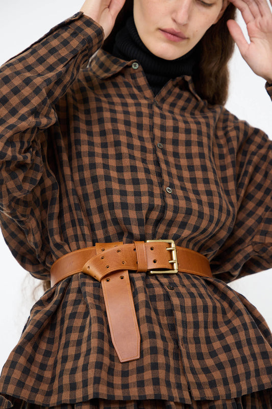 A person wears a brown and black checkered shirt paired with the Leather Belt in Brown from Ichi Antiquités, which features a brass buckle. The individual's head is partly cropped out of the frame.