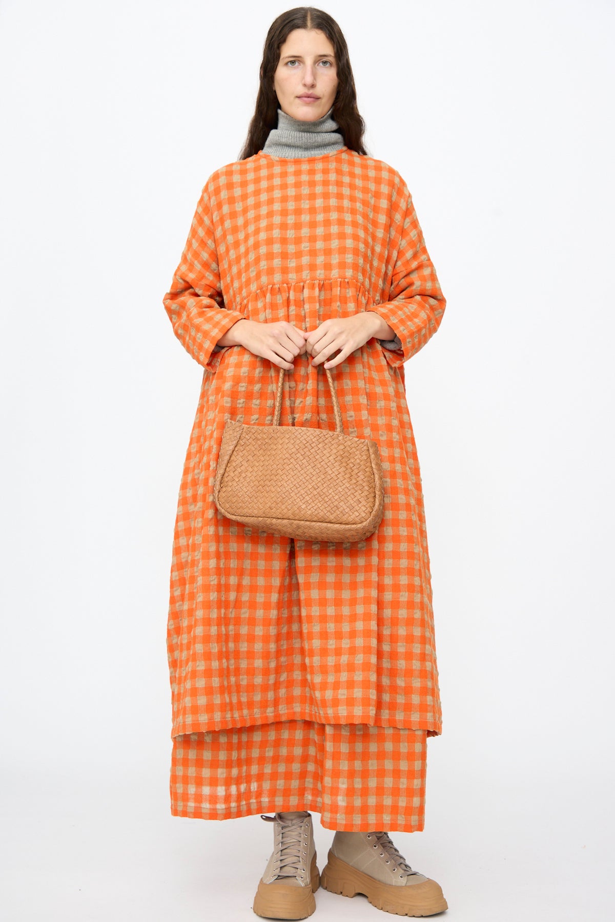 A person wearing a long orange and beige checkered dress and light-colored boots holds a small Ichi Antiquités Leather Tote Bag in Brown.
