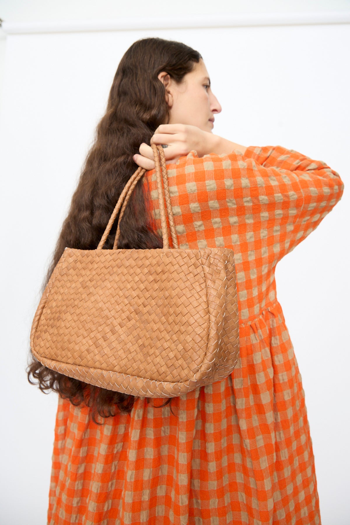 A person wearing an orange gingham dress carries a small, brown Ichi Antiquités Leather Tote Bag over their shoulder.