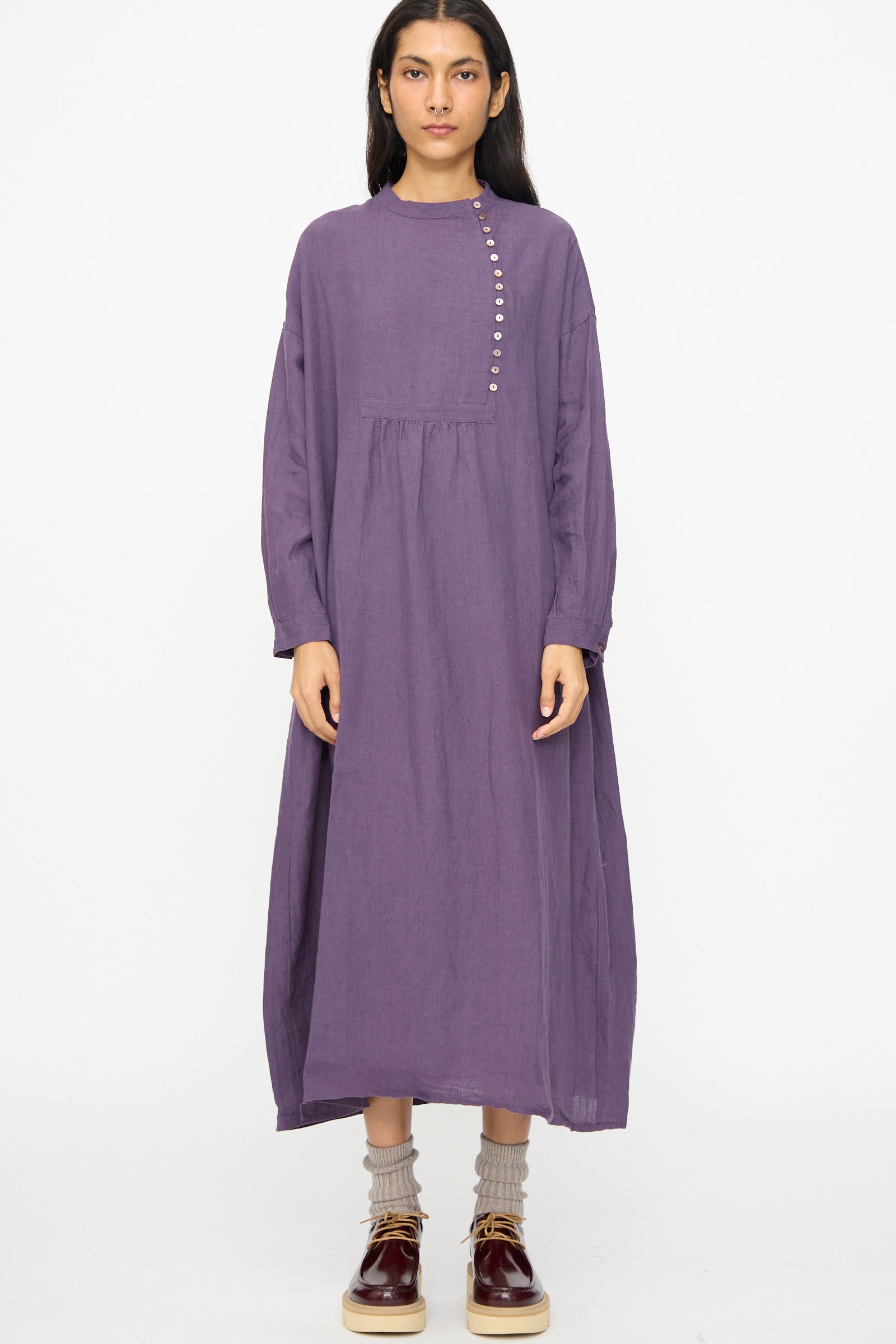 A woman stands facing forward against a plain white background, wearing the Ichi Antiquités Linen Broad Dress in Purple with long sleeves, paired with gray socks and burgundy shoes.