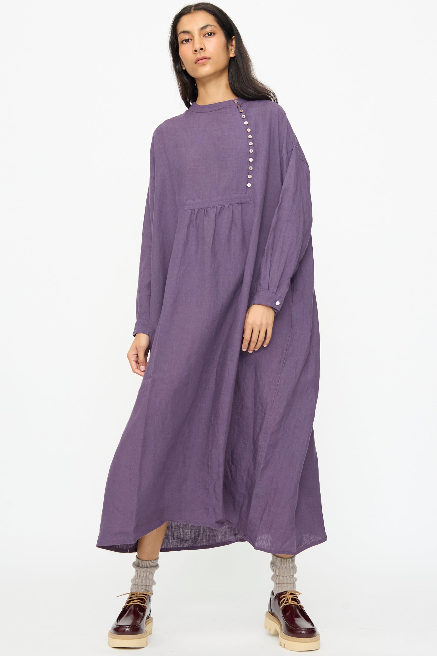 A woman wears the Ichi Antiquités Linen Broad Dress in Purple—made in Japan with long sleeves and front buttons—paired with beige socks and brown platform shoes, set against a plain white background.