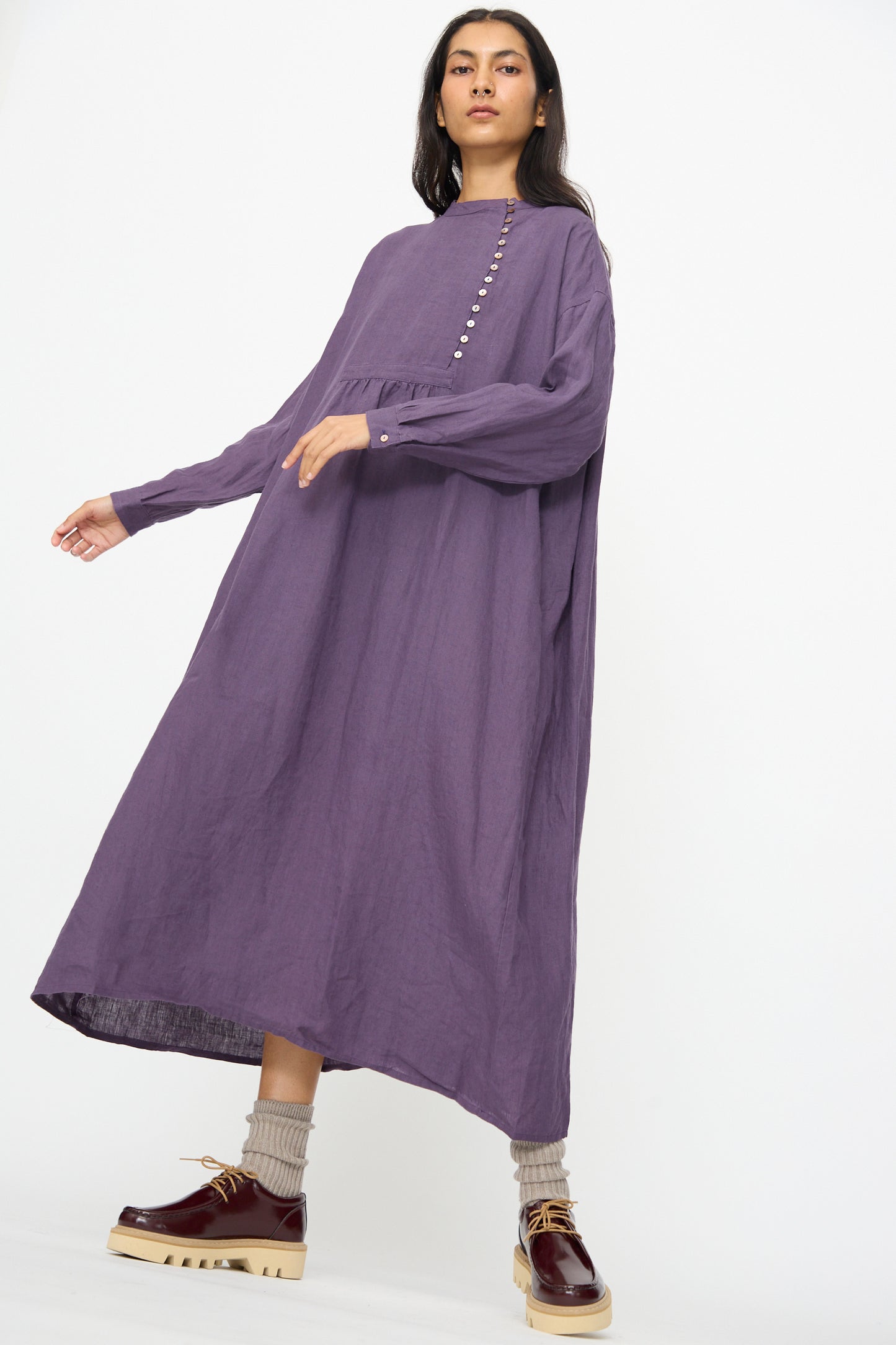 A woman wears the Ichi Antiquités Linen Broad Dress in purple, featuring long sleeves and front buttons, paired with beige socks and brown platform shoes, standing against a plain white background.