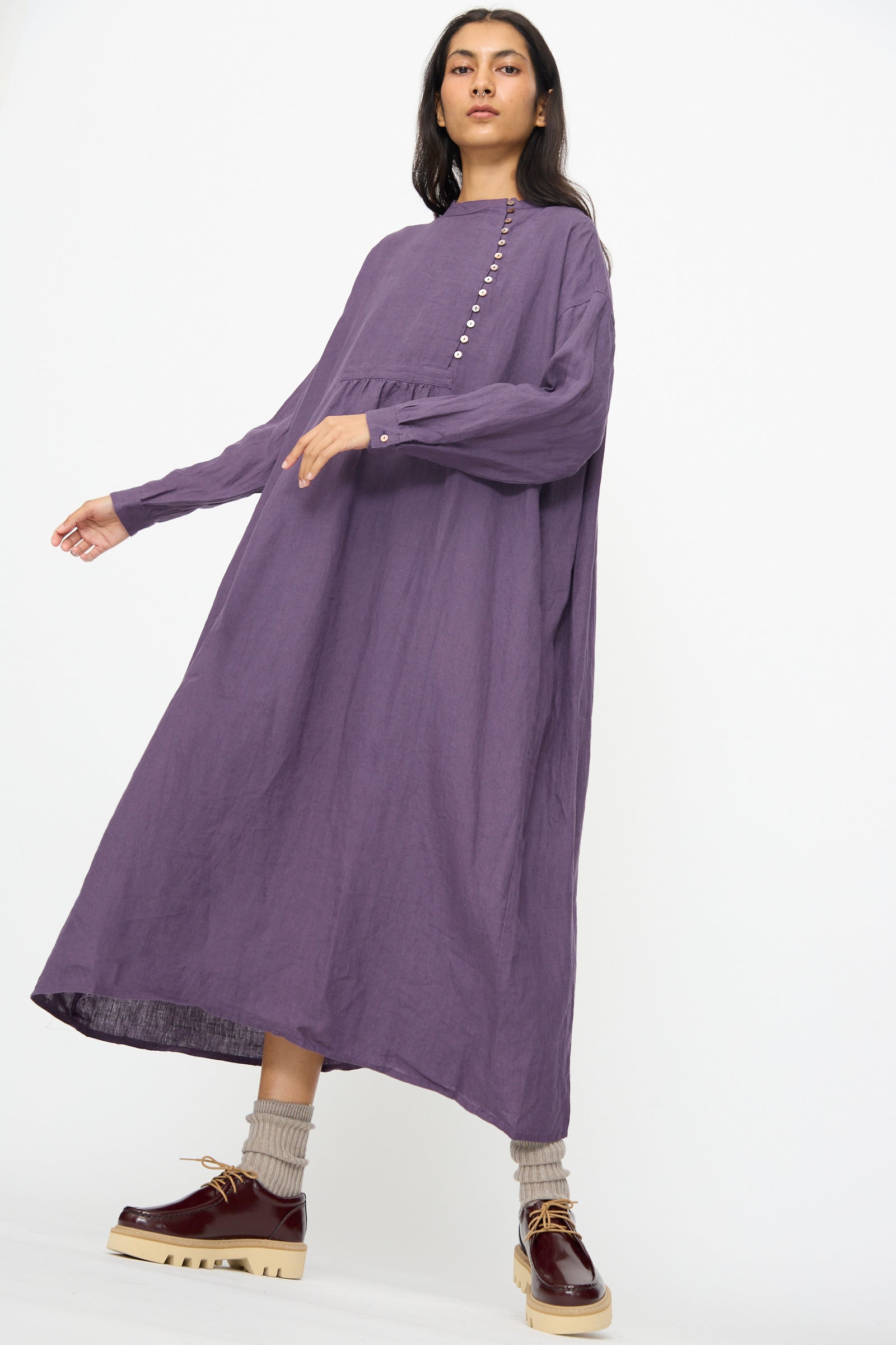 A woman wears the Ichi Antiquités Linen Broad Dress in purple, featuring long sleeves and front buttons, paired with beige socks and brown platform shoes, standing against a plain white background.