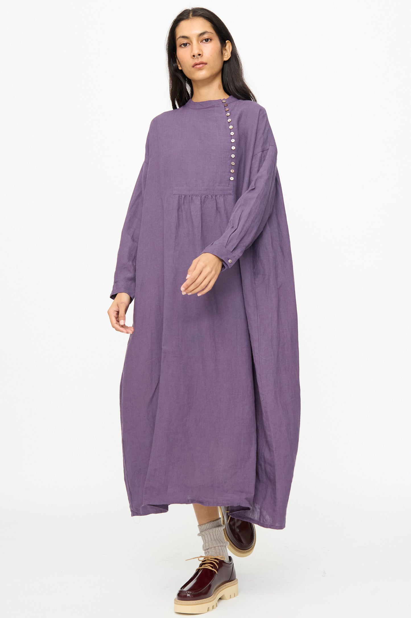 A woman stands against a white background wearing the Ichi Antiquités Linen Broad Dress in Purple, styled with beige socks and burgundy platform shoes.