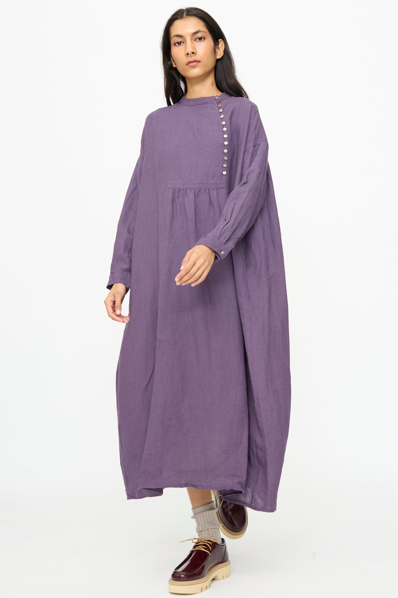 A woman stands against a white background wearing the Ichi Antiquités Linen Broad Dress in Purple, styled with beige socks and burgundy platform shoes.