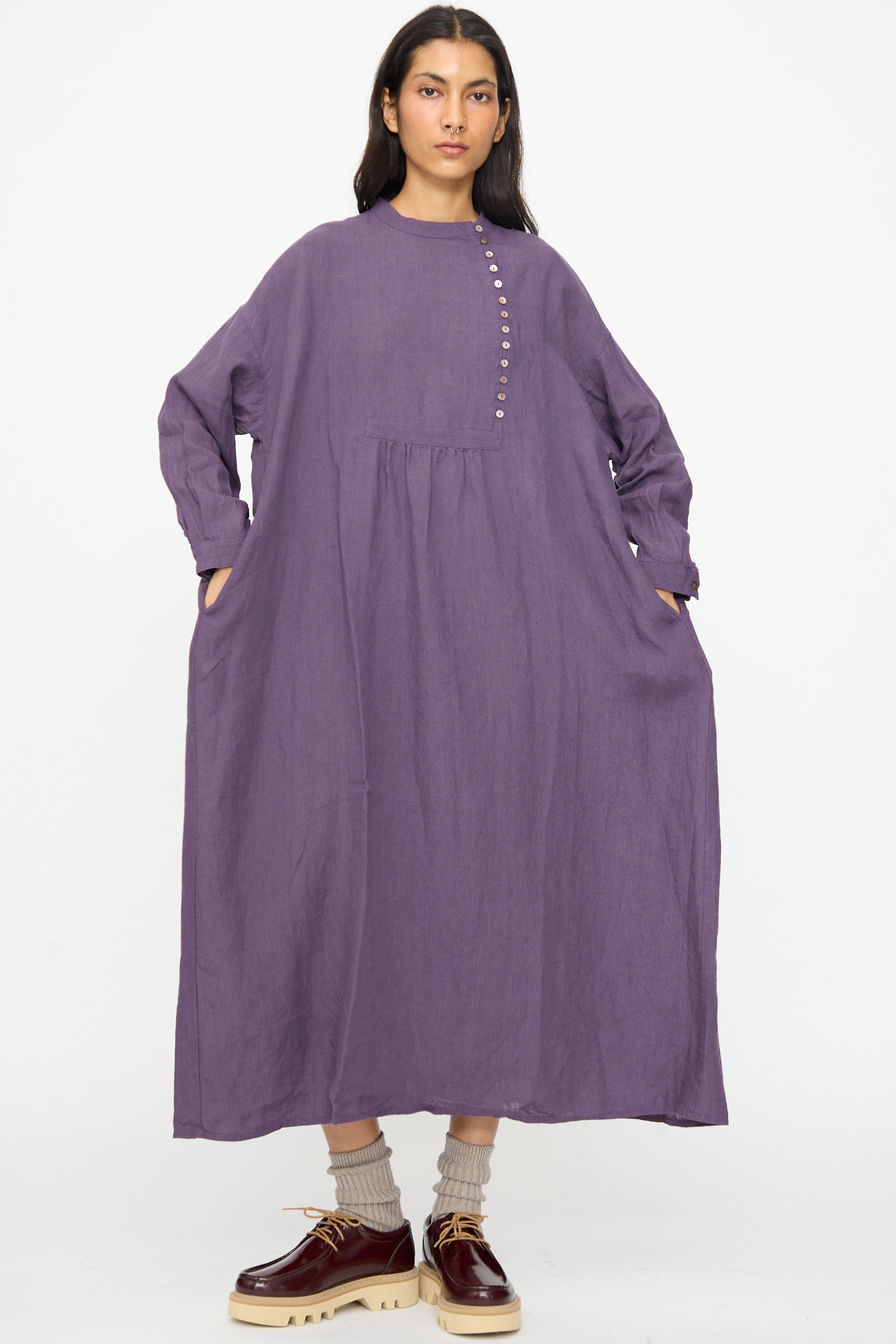 A woman wears the Ichi Antiquités Linen Broad Dress in Purple—loose-fitting with long sleeves, side pockets, and button details—paired with maroon platform shoes and beige socks.