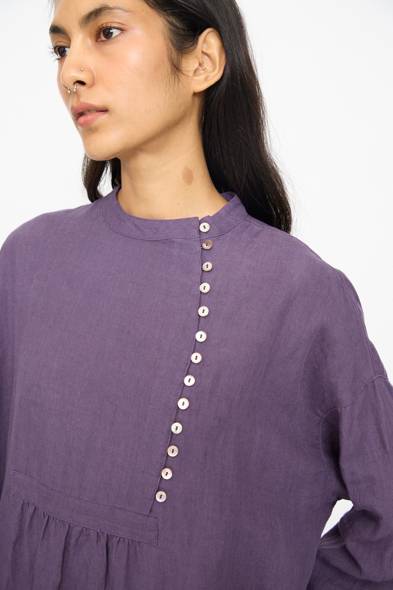 A person wears the Ichi Antiquités Linen Broad Dress in Purple, a long-sleeved Japanese-made maxi with a diagonal button row, standing against a plain, light background.
