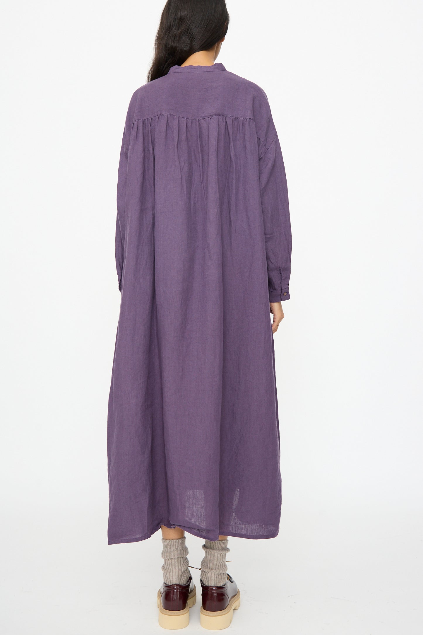 A person stands facing away against a white background, wearing the Ichi Antiquités Linen Broad Dress in purple—a loose-fitting, long-sleeve maxi dress made in Japan—with grey socks and burgundy shoes.