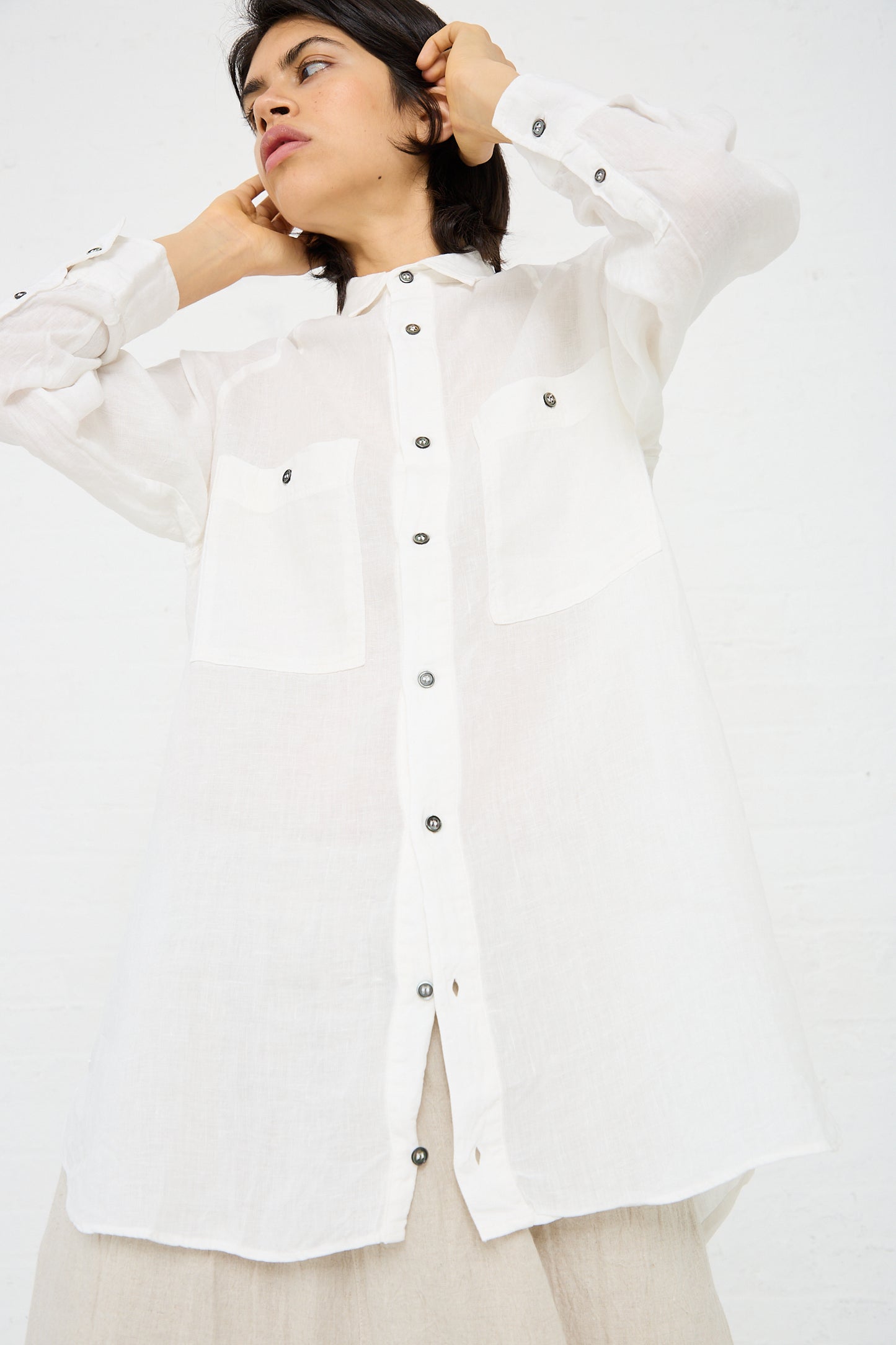 Person wearing a loosely fitted Ichi Antiquités Linen Canvas Overdye Shirt in White with two front pockets, looking to the side, with hands touching the back of their head. Background is plain white.