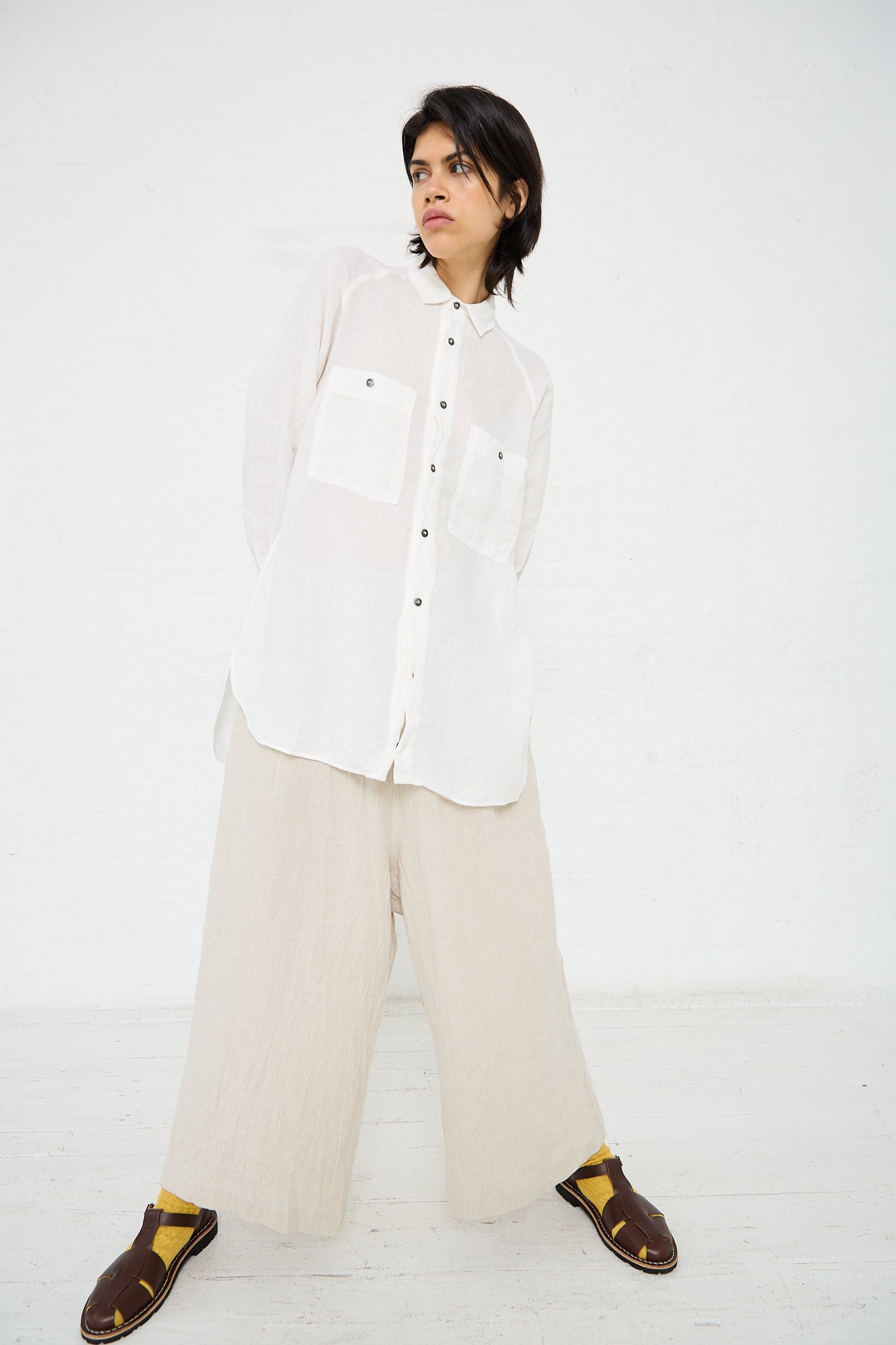 A person stands against a white wall wearing an Ichi Antiquités Linen Canvas Overdye Shirt in White, beige wide-leg pants, yellow socks, and brown sandals. They have short, dark hair and are looking slightly to the side.