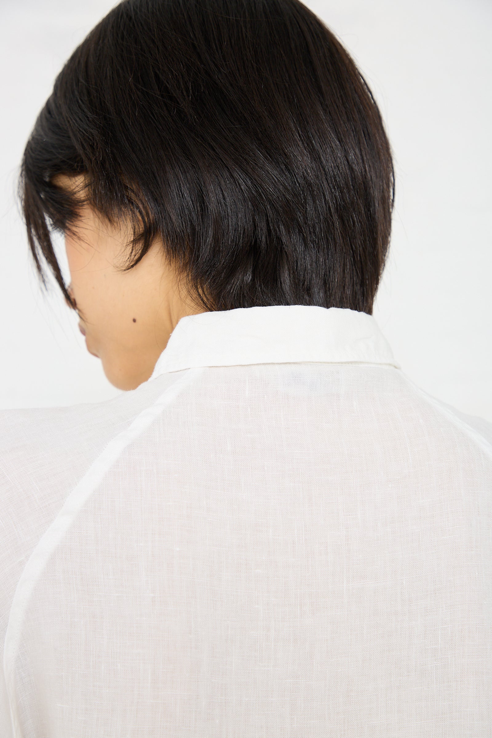 The image shows the back of a person with short, dark hair wearing an oversized fit, white, collared, semi-transparent Linen Canvas Overdye Shirt in White by Ichi Antiquités. The focus is on the shirt's texture and material.