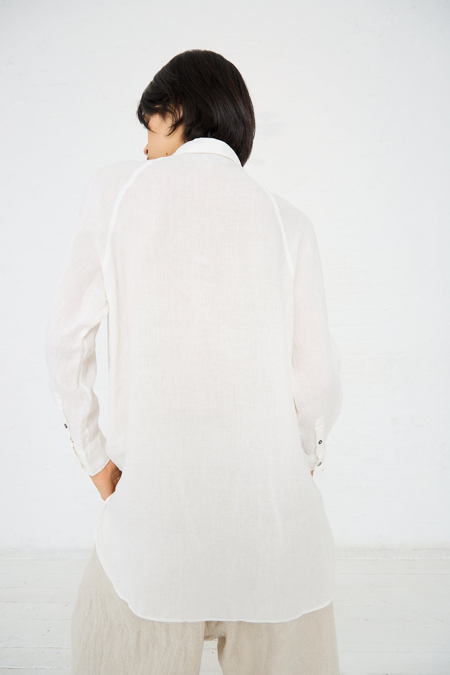 Person with short black hair wearing a loose, long-sleeved Linen Canvas Overdye Shirt in White by Ichi Antiquités and light-colored pants, standing with their back turned.