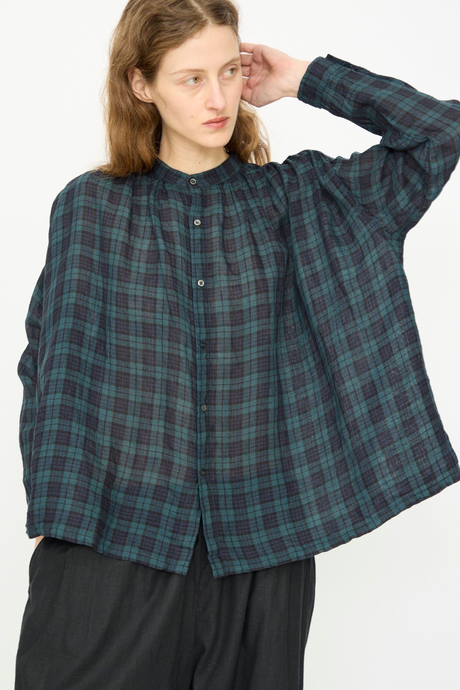 A person with long hair wears the Ichi Antiquités Linen Canvas Shirt in Black Watch, a blue and green plaid oversized linen shirt, with dark pants, standing against a plain white background.