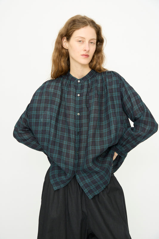 Person with long brown hair wears the Ichi Antiquités Linen Canvas Shirt in Black Watch—a dark green and navy plaid—paired with black pants, standing against a plain white background.