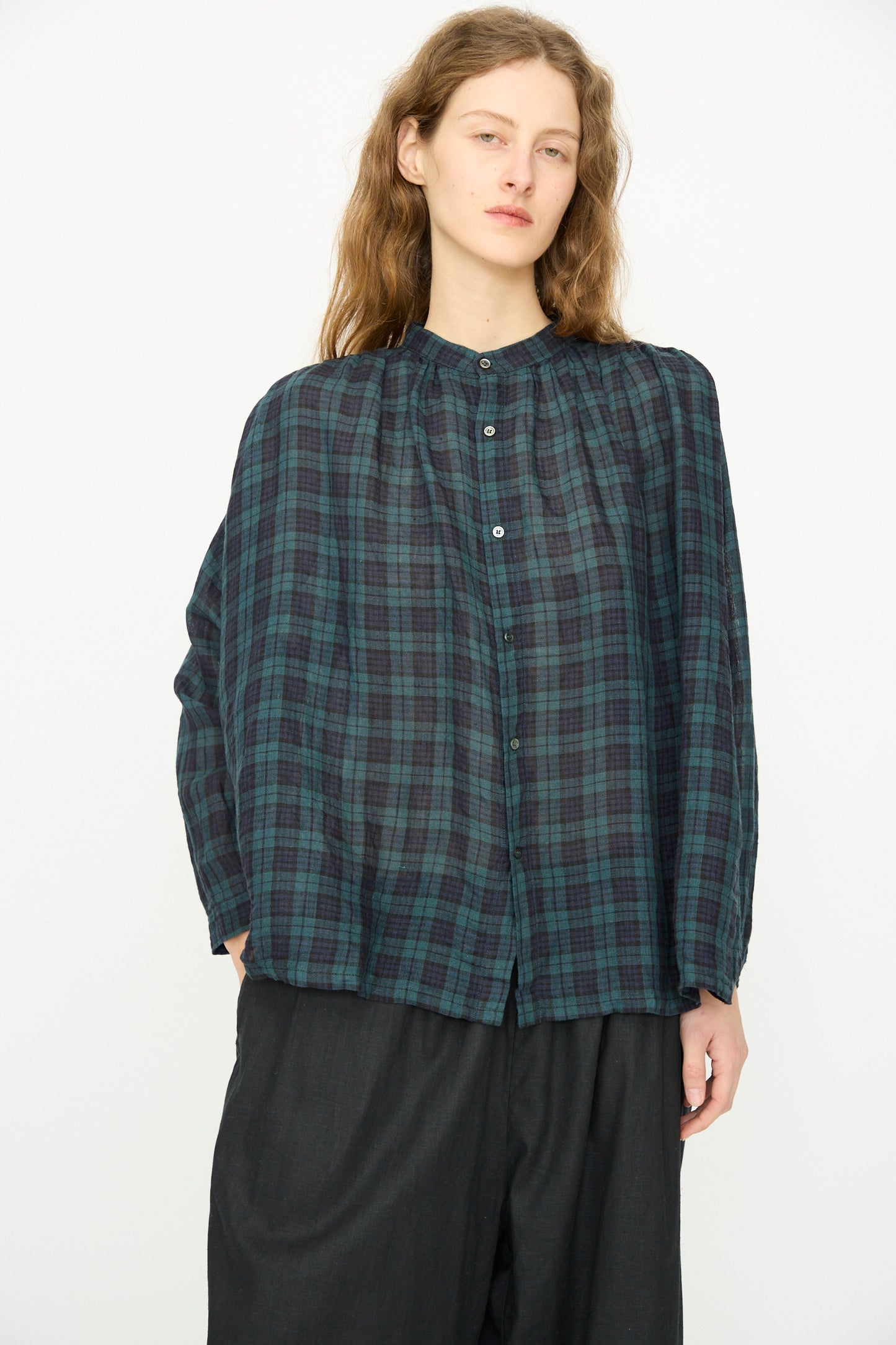 A person with long brown hair wears the Ichi Antiquités Linen Canvas Shirt in Black Watch—its loose, dark green and navy tartan design paired with black pants—standing against a plain white background.