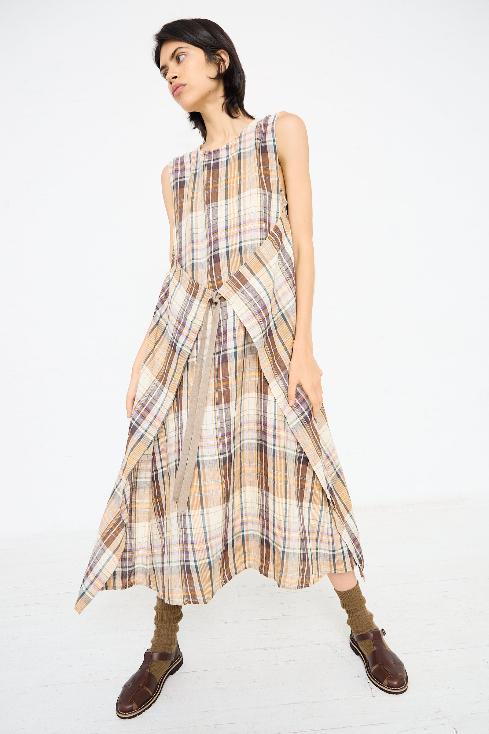 A person stands against a plain background wearing the Ichi Antiquités Linen Check Dress in Beige, a sleeveless, multicolor linen dress with an asymmetrical hem and brown shoes. The wrap detail of the dress includes shades of beige, blue, and red.