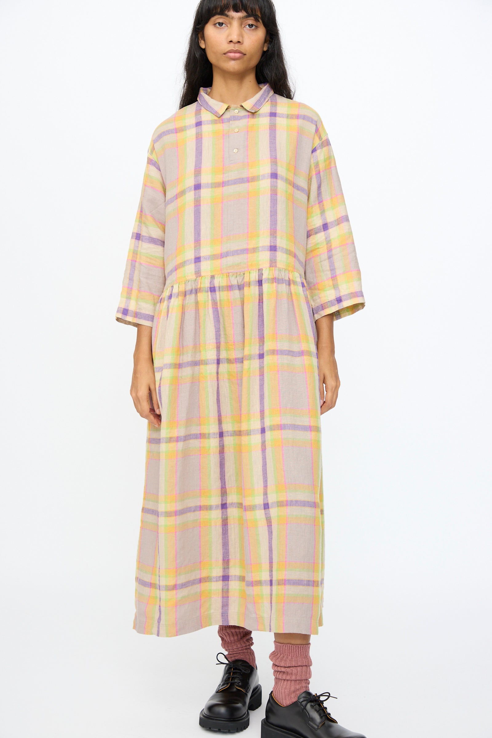 A person wearing the Ichi Antiquités Linen Check Dress in Beige stands against a plain white background, paired with pink socks and black shoes.