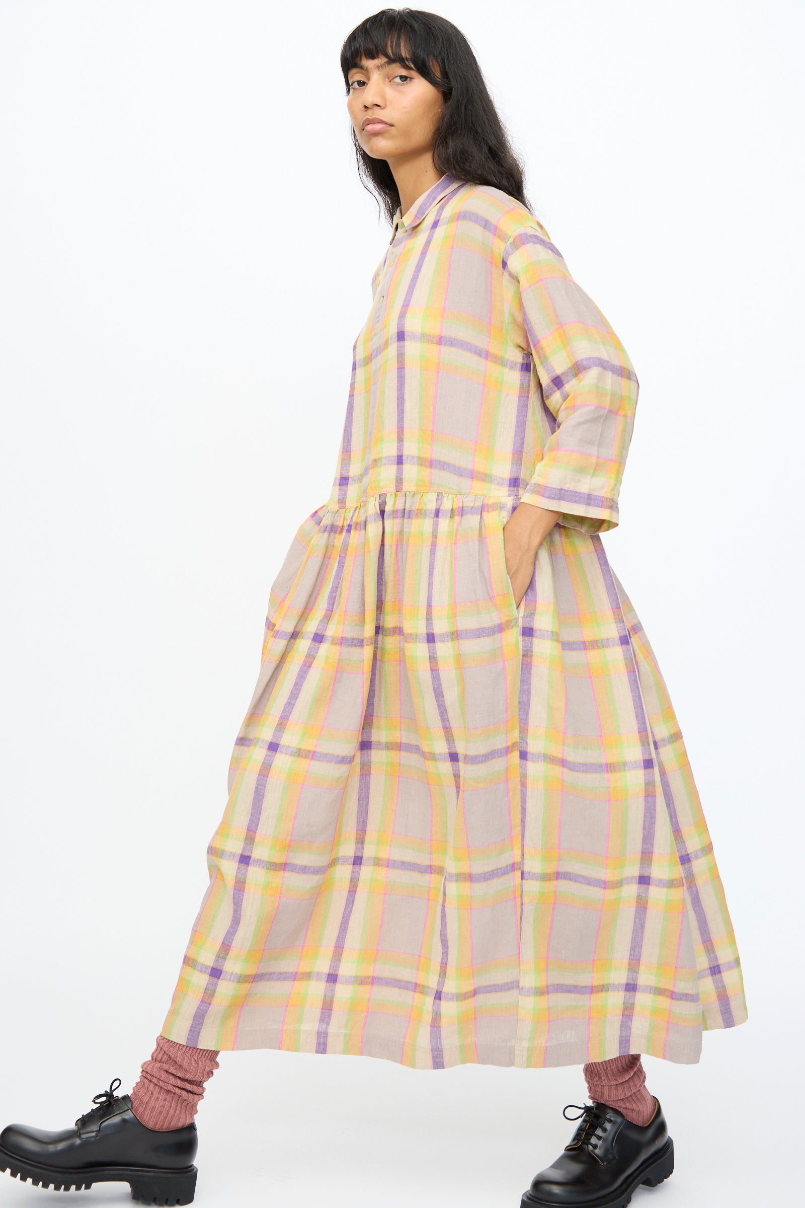 A person wears the Ichi Antiquités Linen Check Dress in Beige, an oversized Japanese-made piece, styled with pink socks and black lace-up shoes, walking against a plain white background.