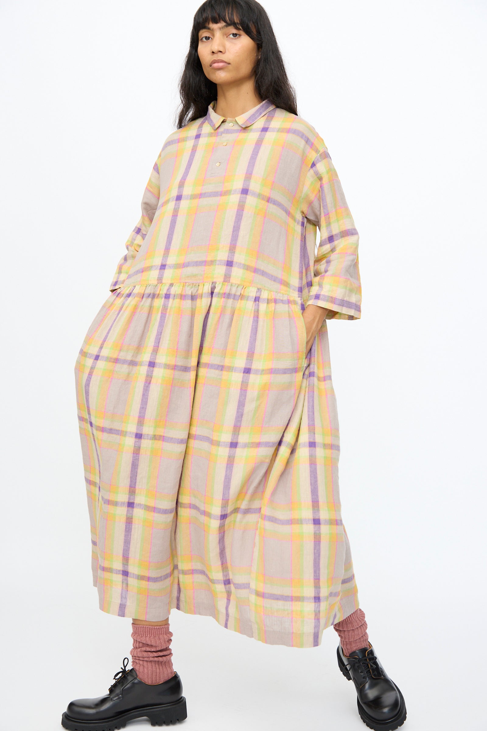 A woman stands with hands in pockets against a white background, wearing the Ichi Antiquités Linen Check Dress in Beige, paired with pink socks and black shoes.