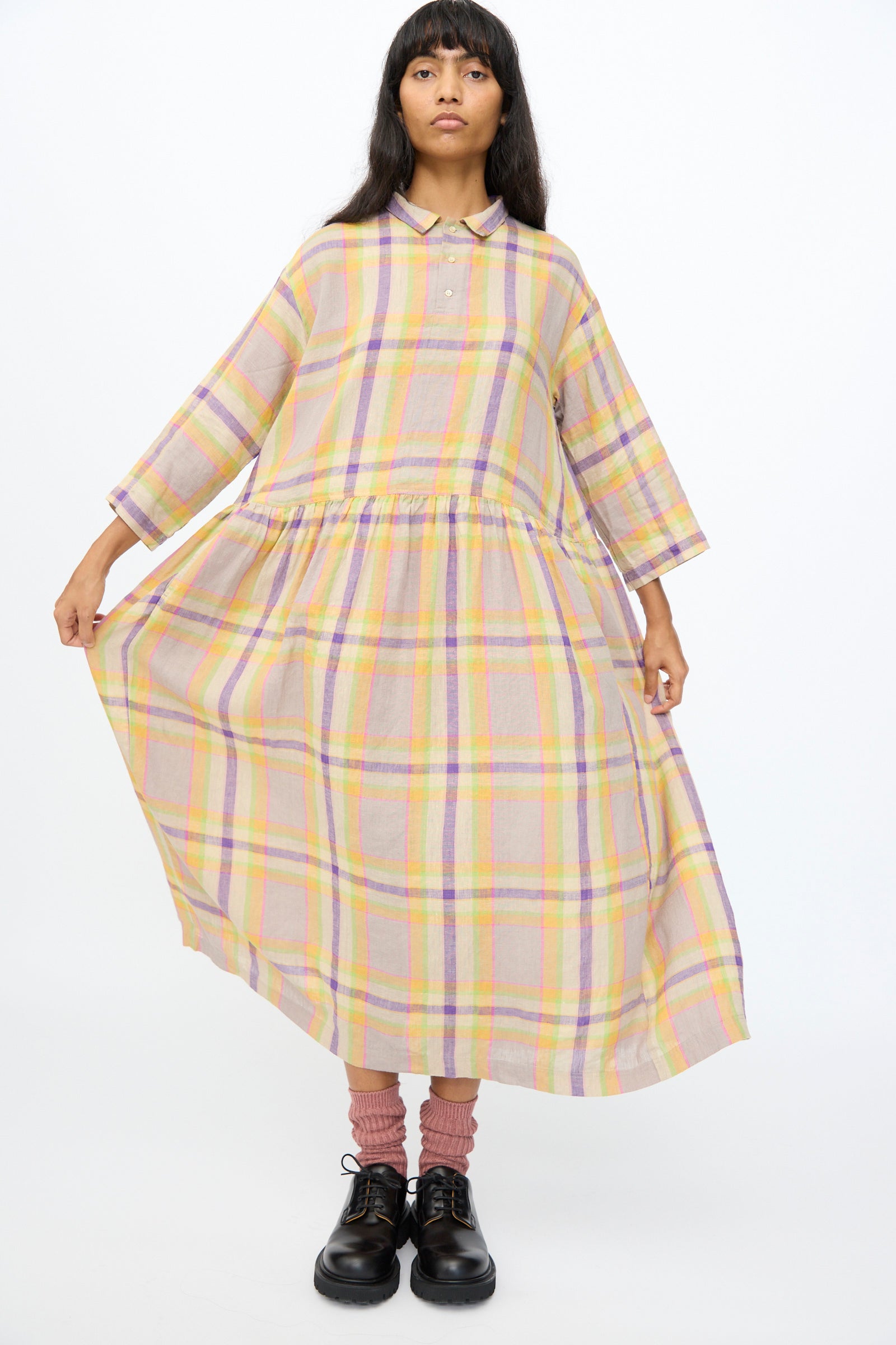 A woman wears the Ichi Antiquités Linen Check Dress in Beige—an oversized linen piece—paired with black shoes and pink socks, standing against a plain white background.