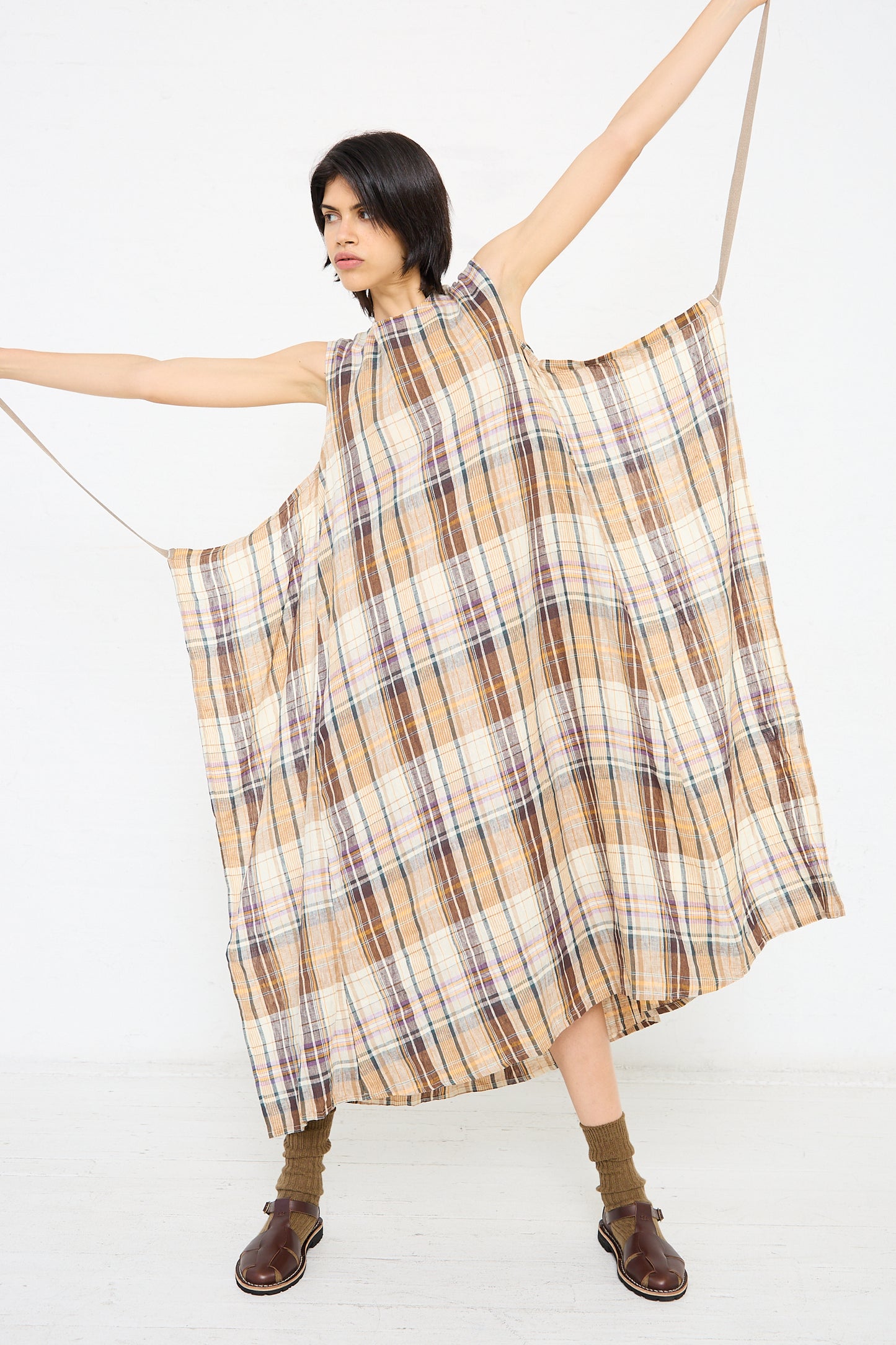 A person stands with arms outstretched, wearing a loose, sleeveless Linen Check Dress in Beige from Ichi Antiquités, brown socks, and brown shoes against a plain white background.