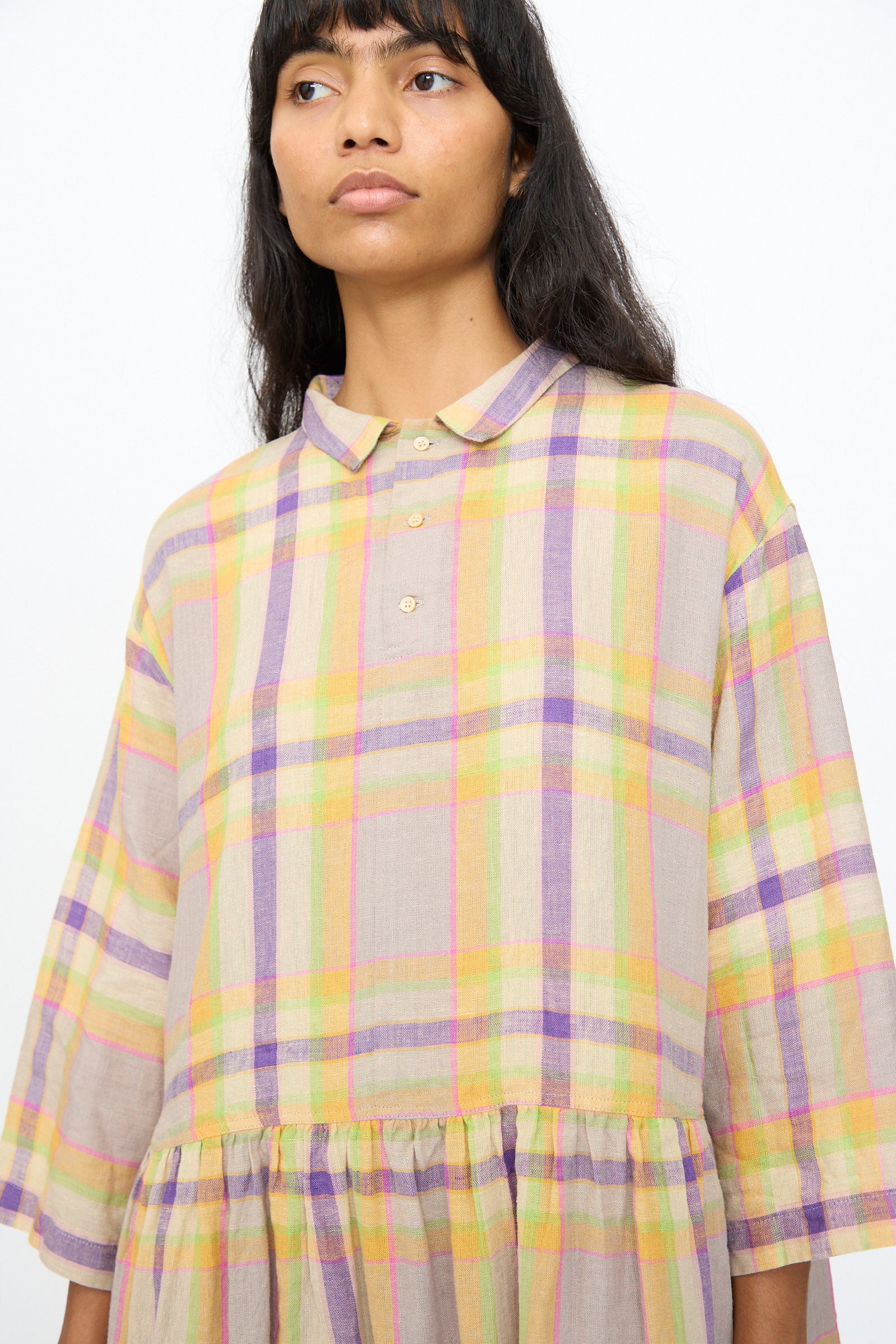 A person wears the Ichi Antiquités Linen Check Dress in Beige—an oversized linen dress with pastel plaid stripes, collar, and front buttons against a white backdrop. 