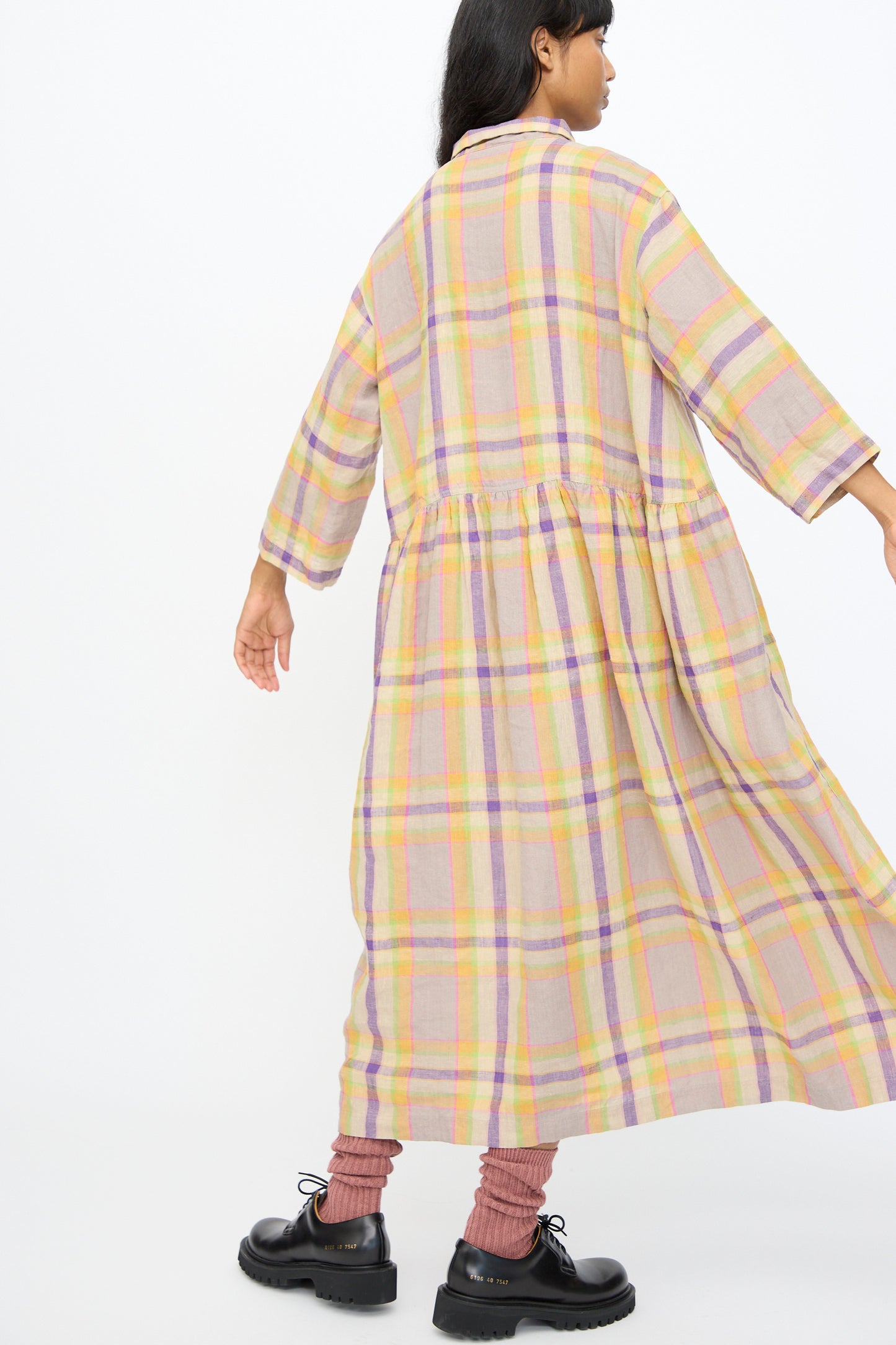 A woman wearing the Ichi Antiquités Linen Check Dress in Beige stands against a plain white background, facing away. This oversized linen dress is made in Japan. She pairs it with pink socks and black shoes.