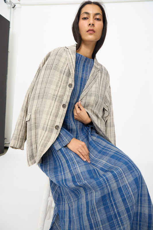 A woman sits on a stool against a plain white background, wearing the Ichi Antiquités Linen Check Jacket in Natural over a blue plaid dress.