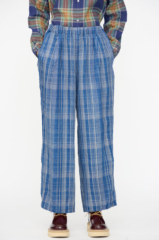 A person stands with hands in pockets, wearing Ichi Antiquités' Linen Check Pants in Navy Indigo, a multicolored plaid shirt, and burgundy platform shoes against a plain background.