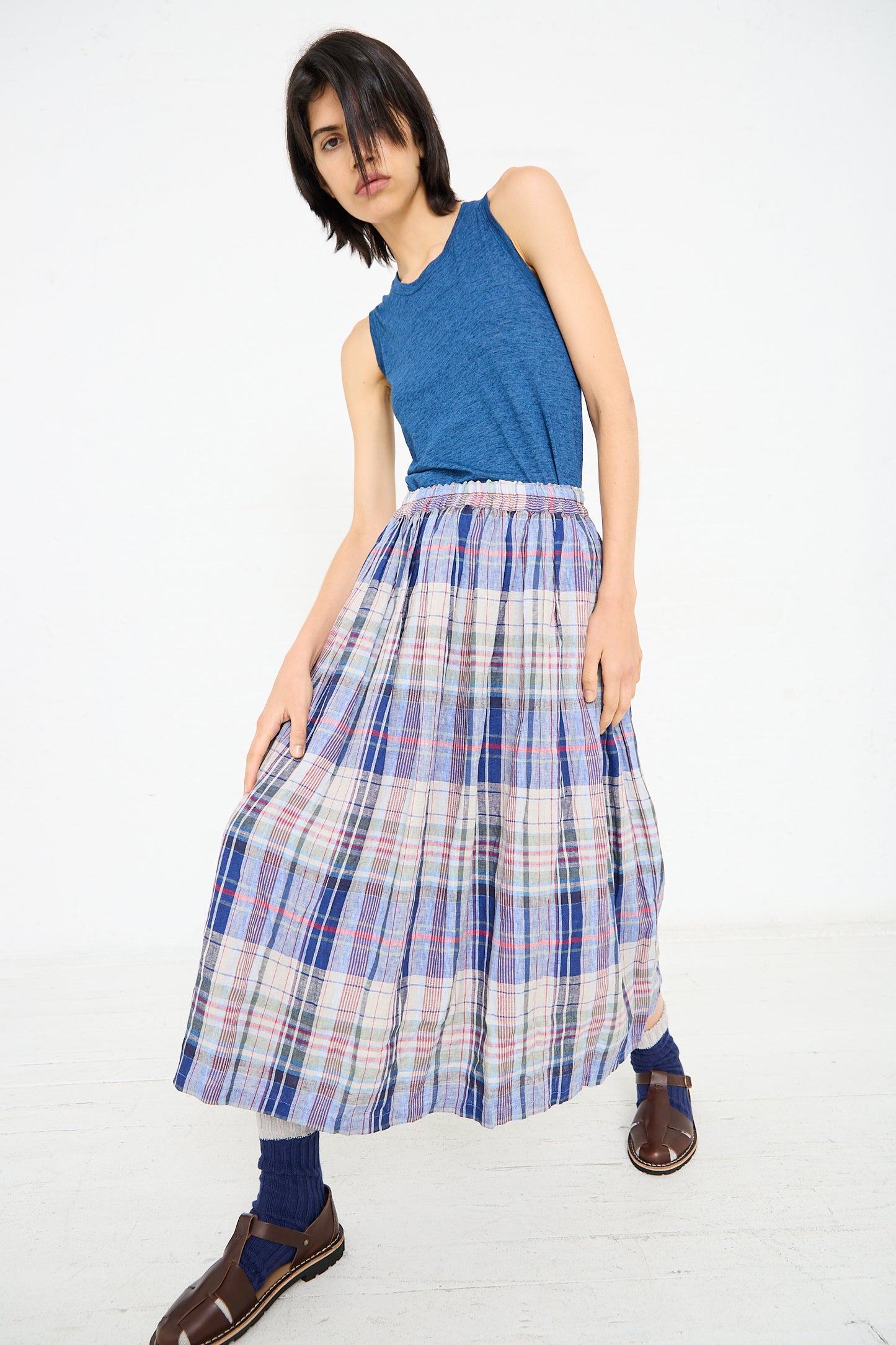 A person with short dark hair wears a blue sleeveless top, a Linen Check Skirt in Navy by Ichi Antiquités, blue socks, and brown sandals, posing against a plain white background.