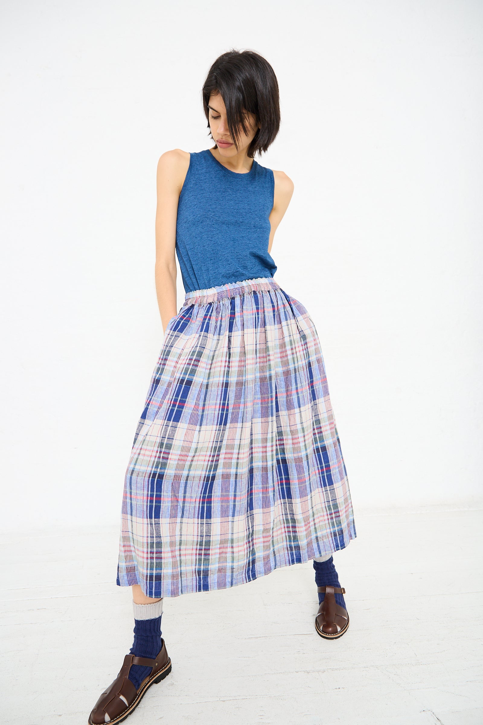 A person stands against a plain background wearing a blue sleeveless top paired with the Linen Check Skirt in Navy by Ichi Antiquités, brown sandals, and blue socks.