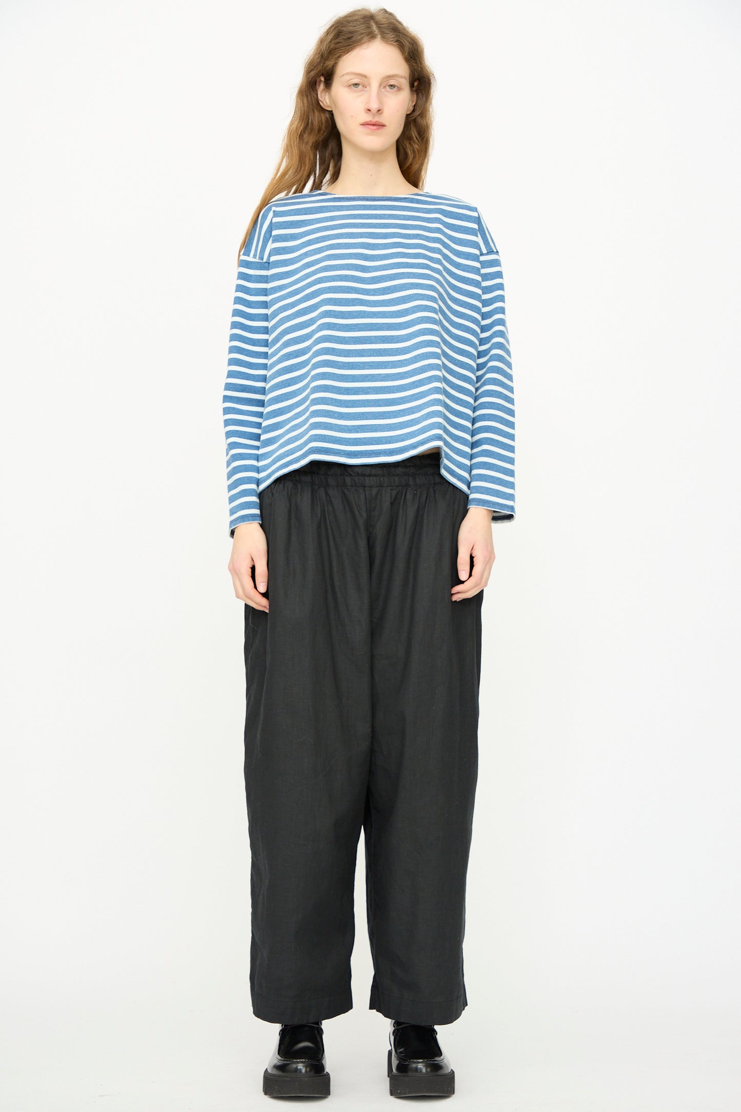 A person stands facing forward against a plain white background, wearing the Linen Cotton Canvas Pant in Black by Ichi Antiquités, paired with a blue and white striped long-sleeve shirt and black shoes.
