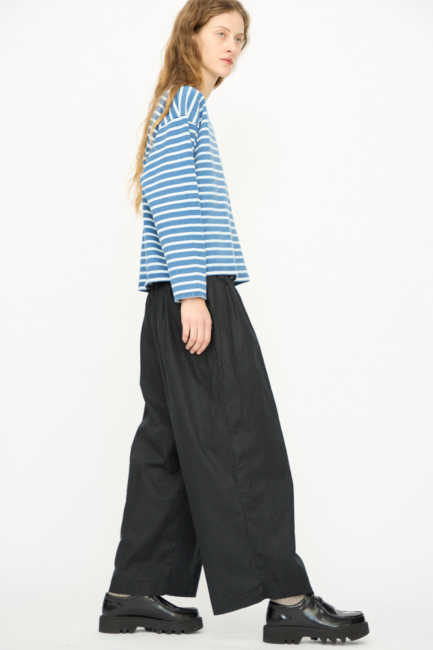 A person stands sideways against a white background, wearing an Ichi Antiquités blue and white striped shirt, Linen Cotton Canvas Pant in Black, and black platform shoes.