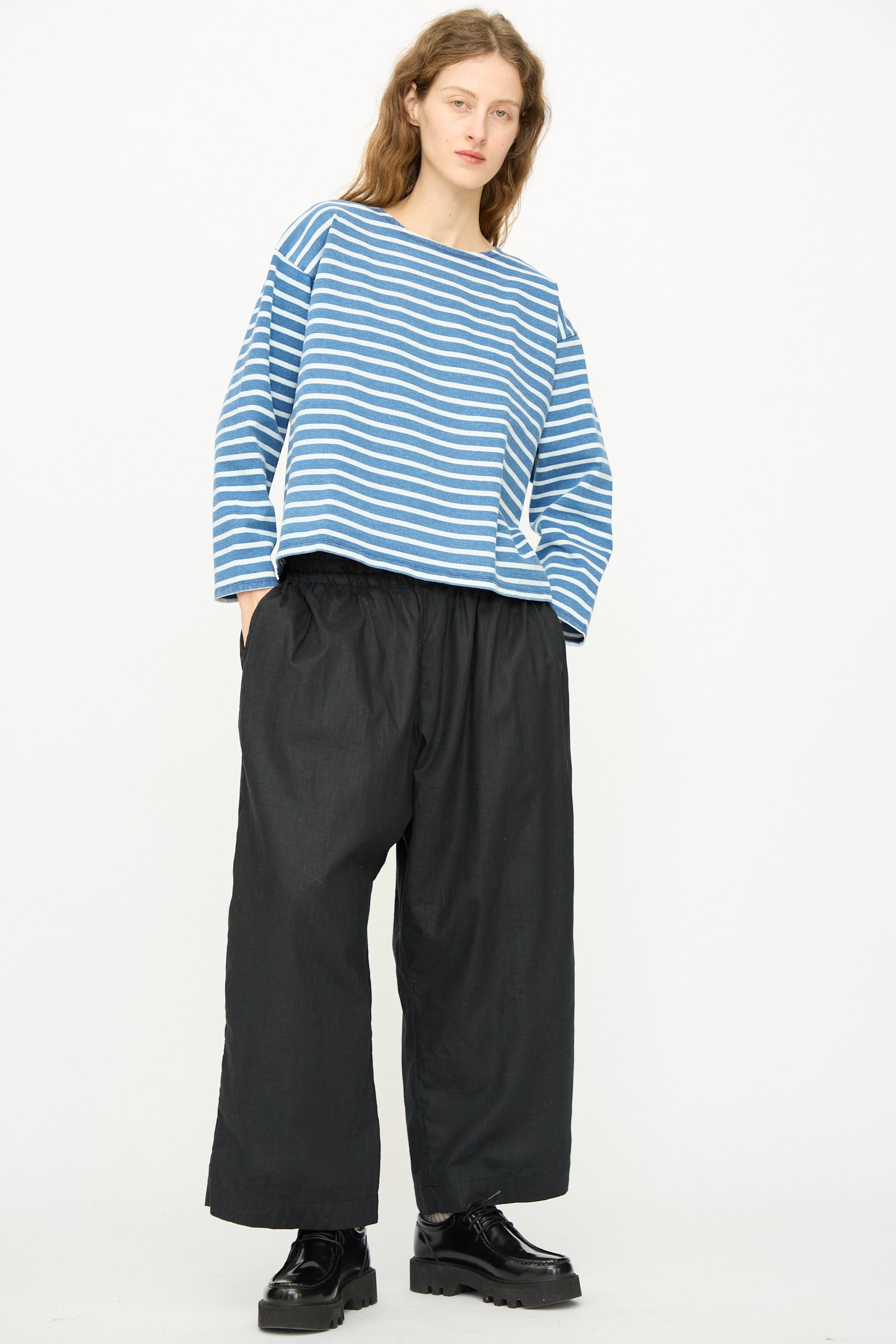 A person stands against a plain white background wearing a blue and white striped long-sleeve shirt, Ichi Antiquités Linen Cotton Canvas Pant in Black, and black shoes.
