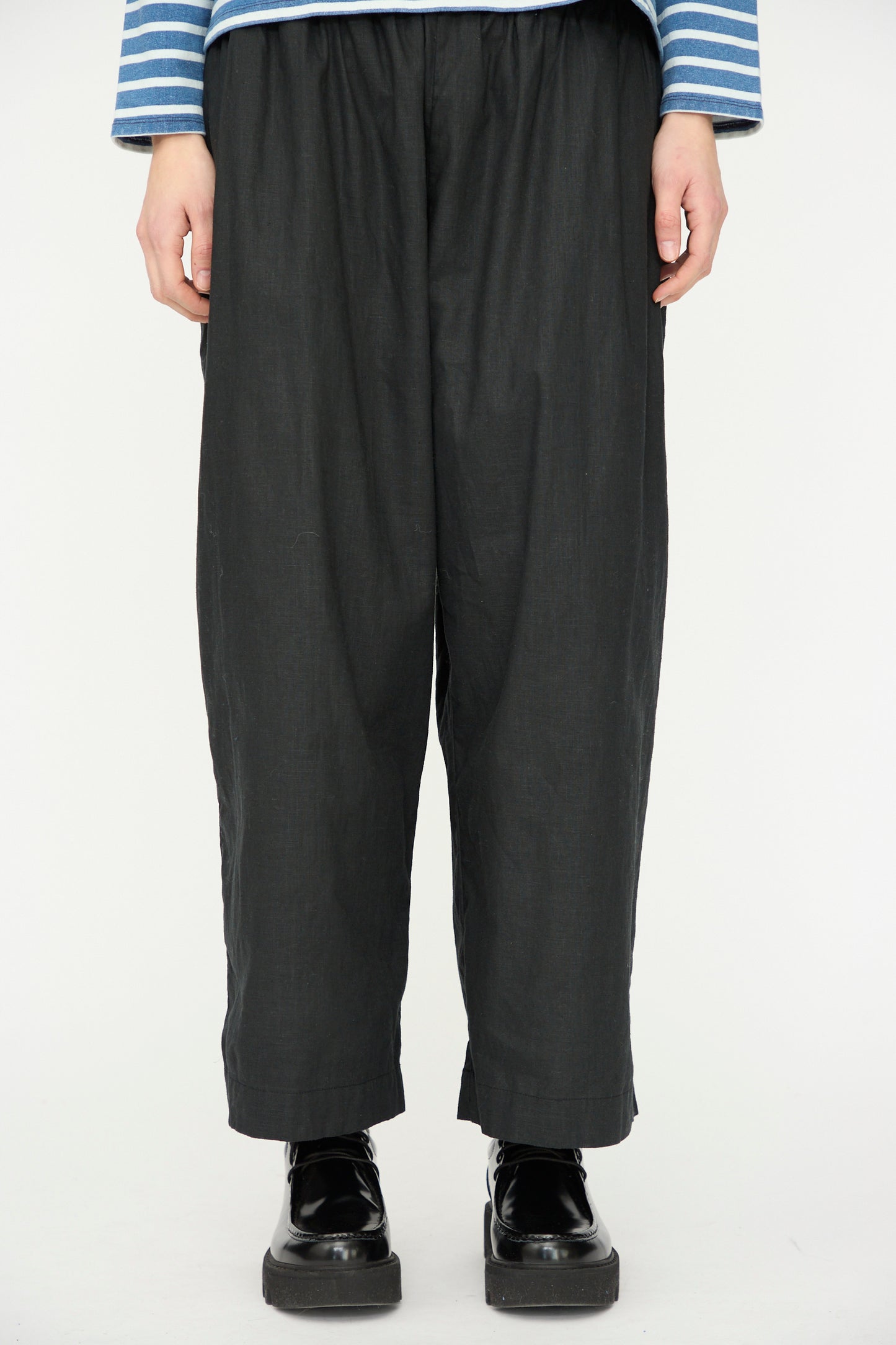 A person wearing the Ichi Antiquités Linen Cotton Canvas Pant in Black, paired with a blue and white striped long-sleeve shirt and black platform shoes, stands against a plain white background.